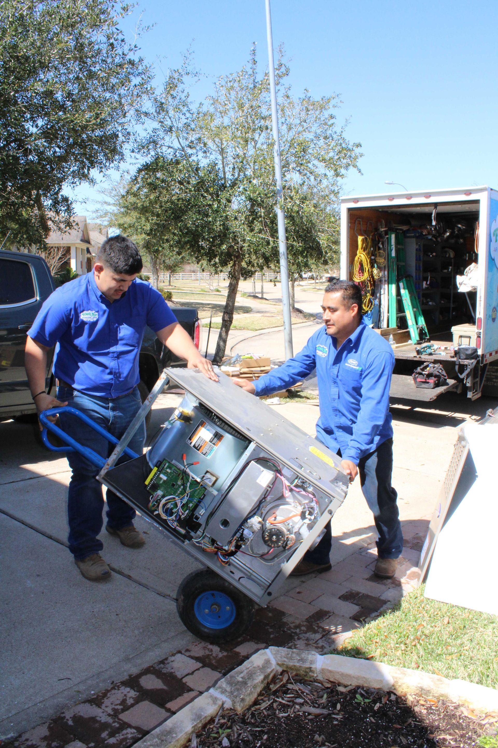 Two men in blue shirts unloading HVAC unit from a truck. Sunny day, residential driveway.
