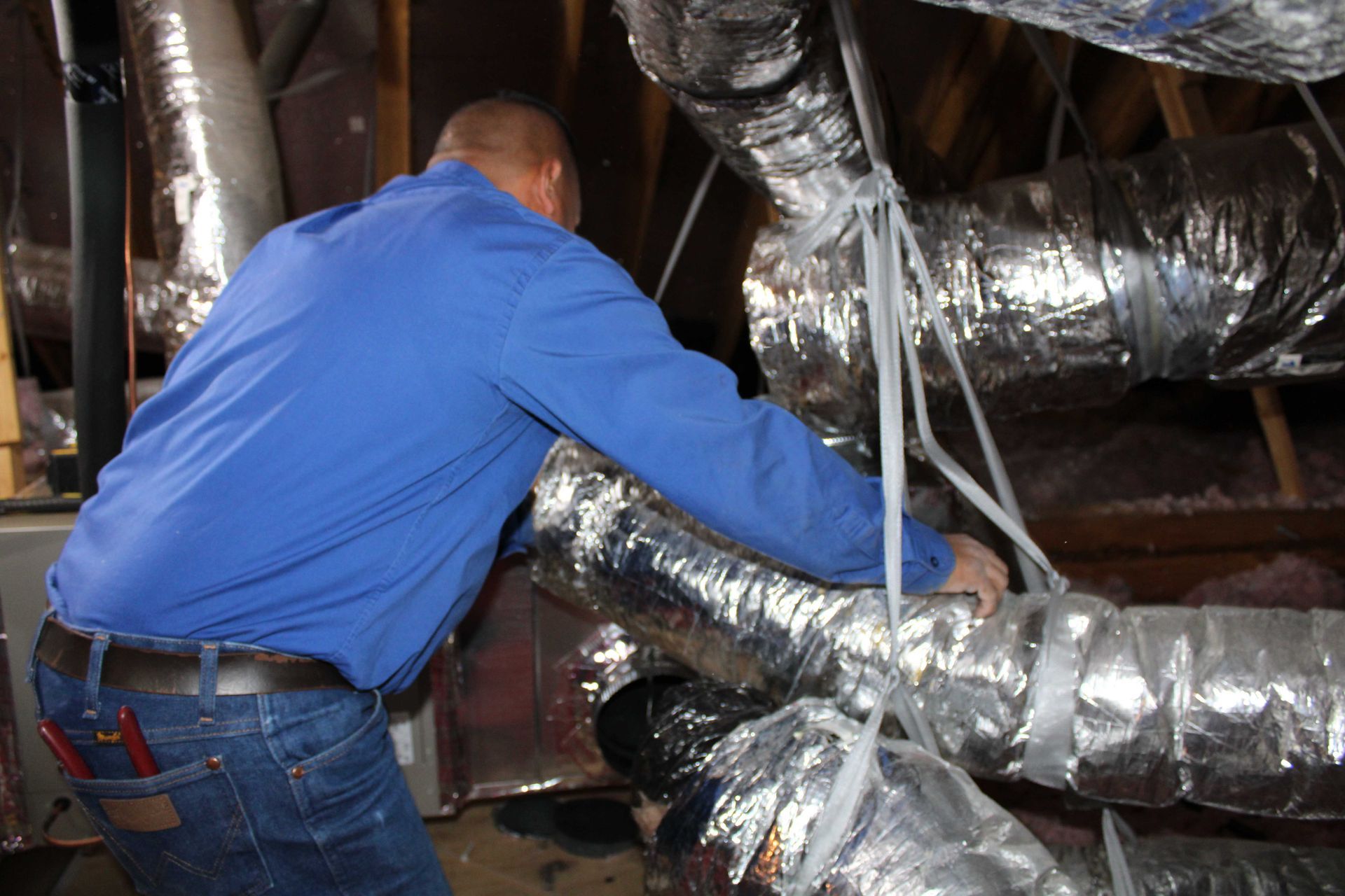 A person in blue shirt working on silver HVAC ductwork in an attic.
