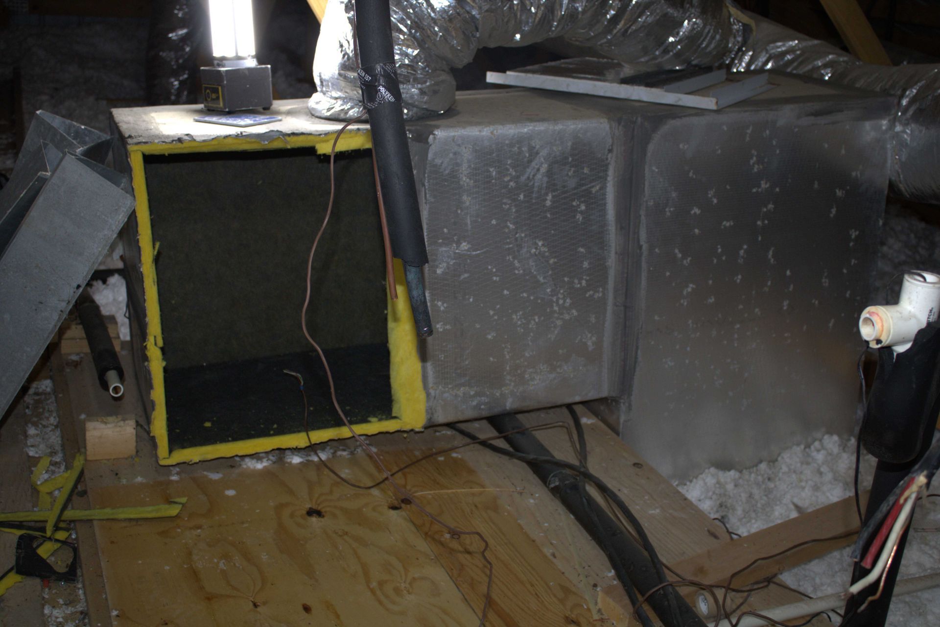 HVAC system in an attic. Ductwork, filter box, and insulation on wooden flooring. Yellow and silver colors.