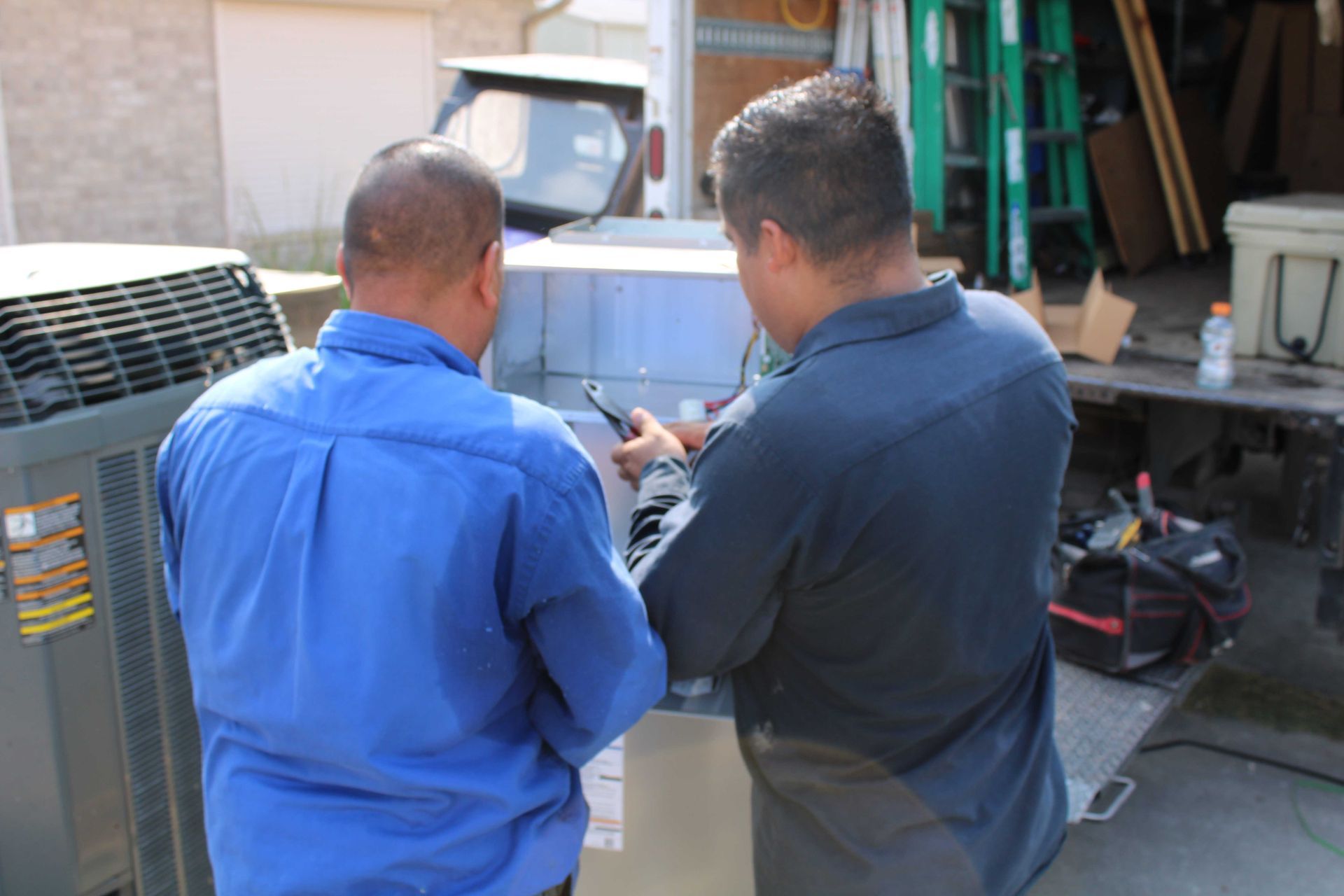 Two technicians working on an AC unit outside. One in blue, one in dark blue, using tools.