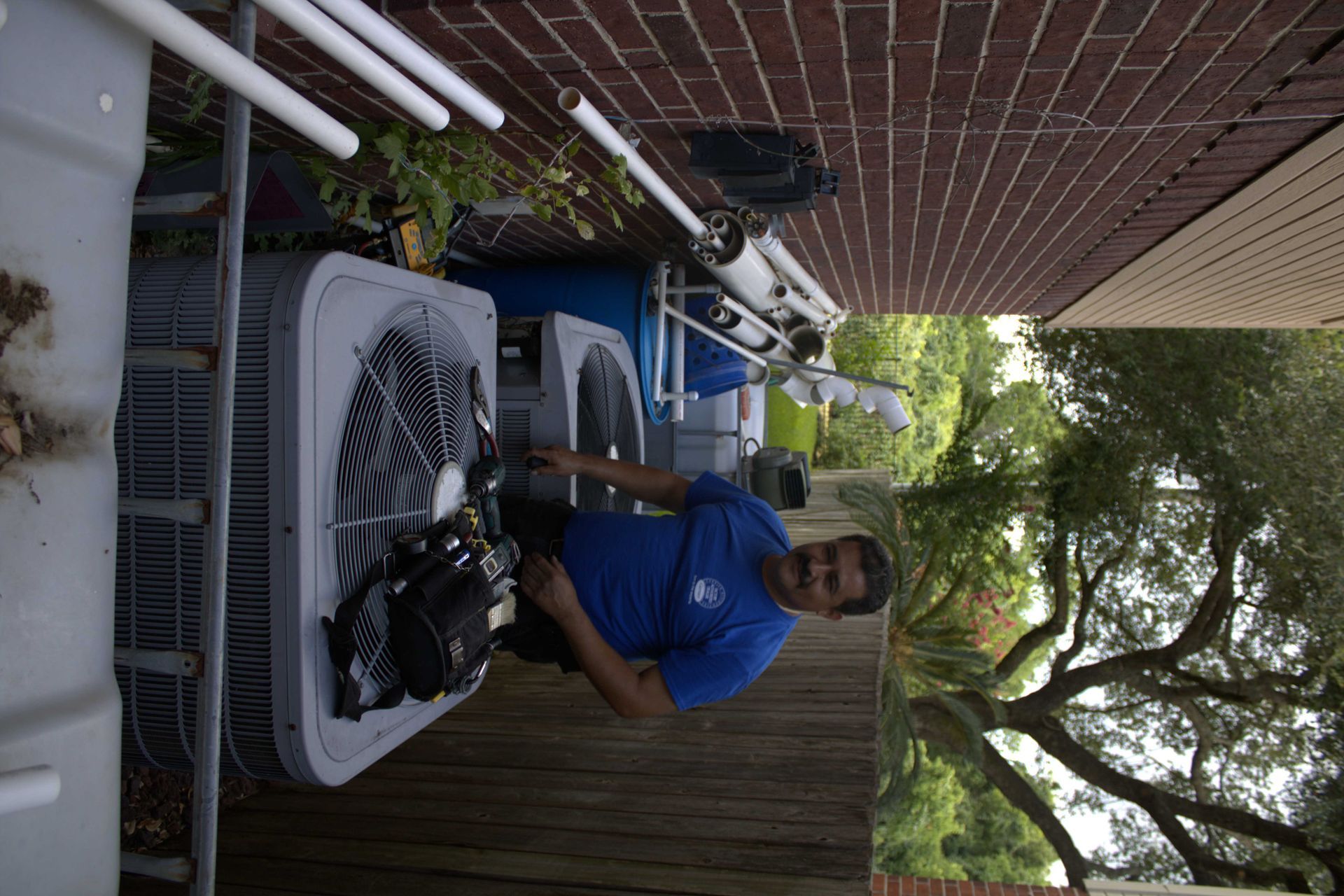 Man in blue shirt working on AC units outdoors next to brick building.