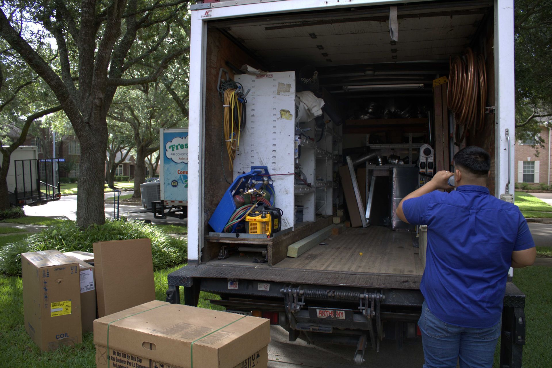A person drinks water near an open moving truck filled with appliances and boxes on a sunny day.