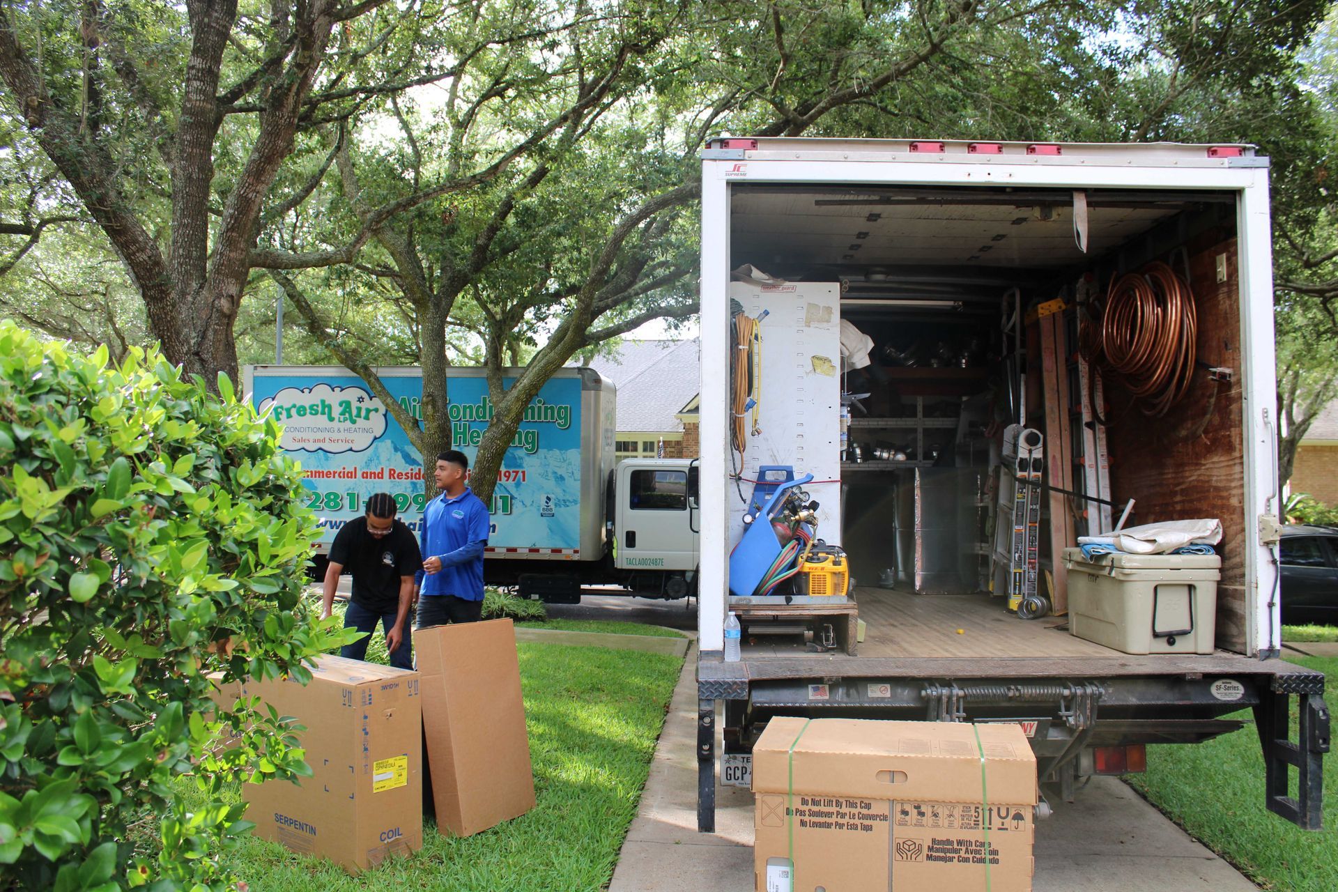 Two moving trucks in driveway; workers load boxes. Boxes on lawn, trees and houses in background.