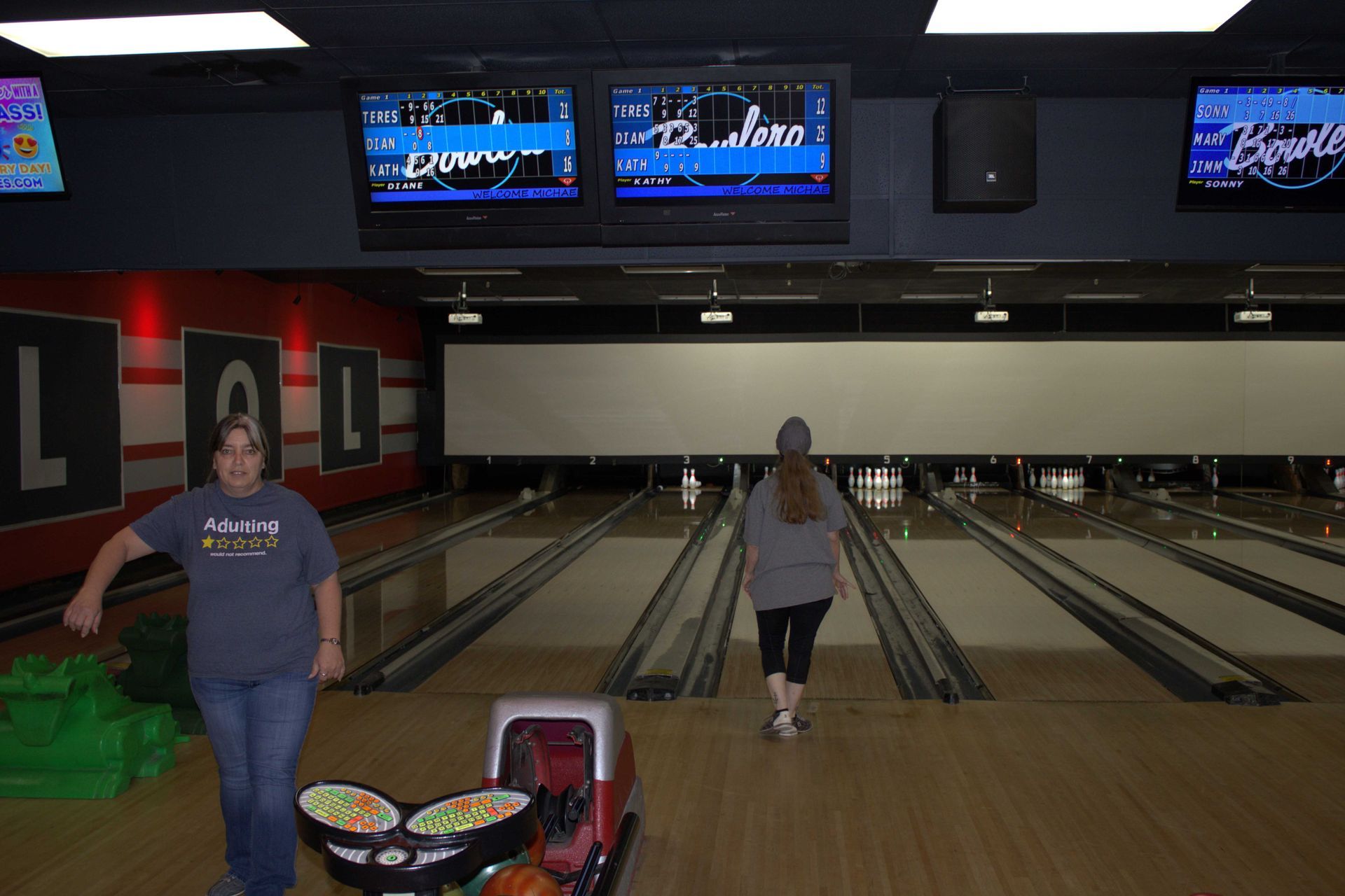 Two women at a bowling alley. One near balls, the other walking towards the lanes. Overhead TVs display scores.