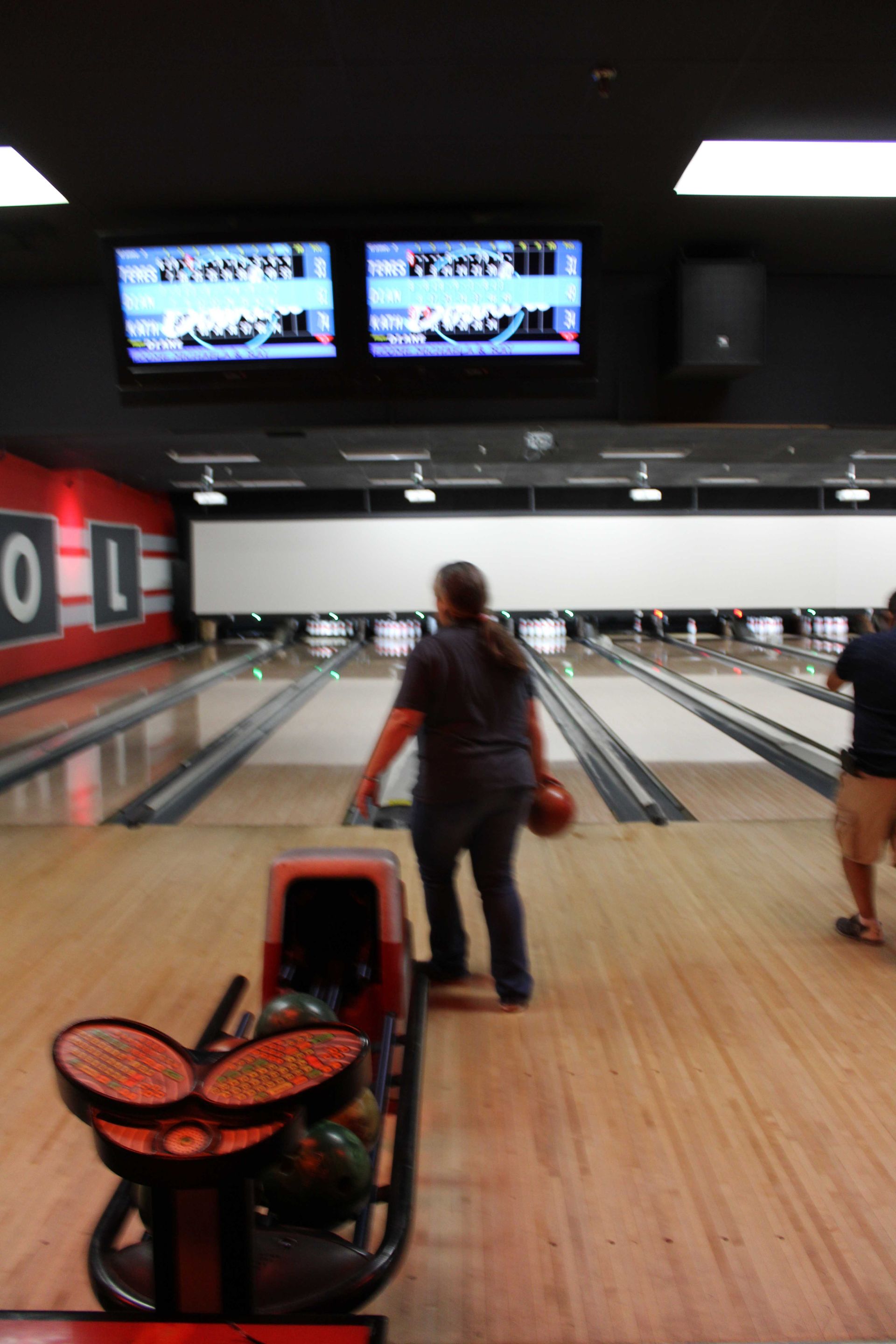 Person bowling, holding ball, approaching lane. Bowling alley interior, scoreboard visible.