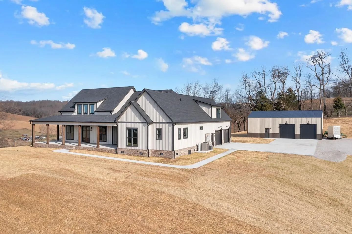 Modern white farmhouse with black roof and detached garage on a large gravel lot under a blue sky