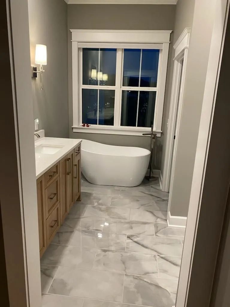 Modern bathroom with gray marble floor, wood vanity, and a white freestanding tub by a window.