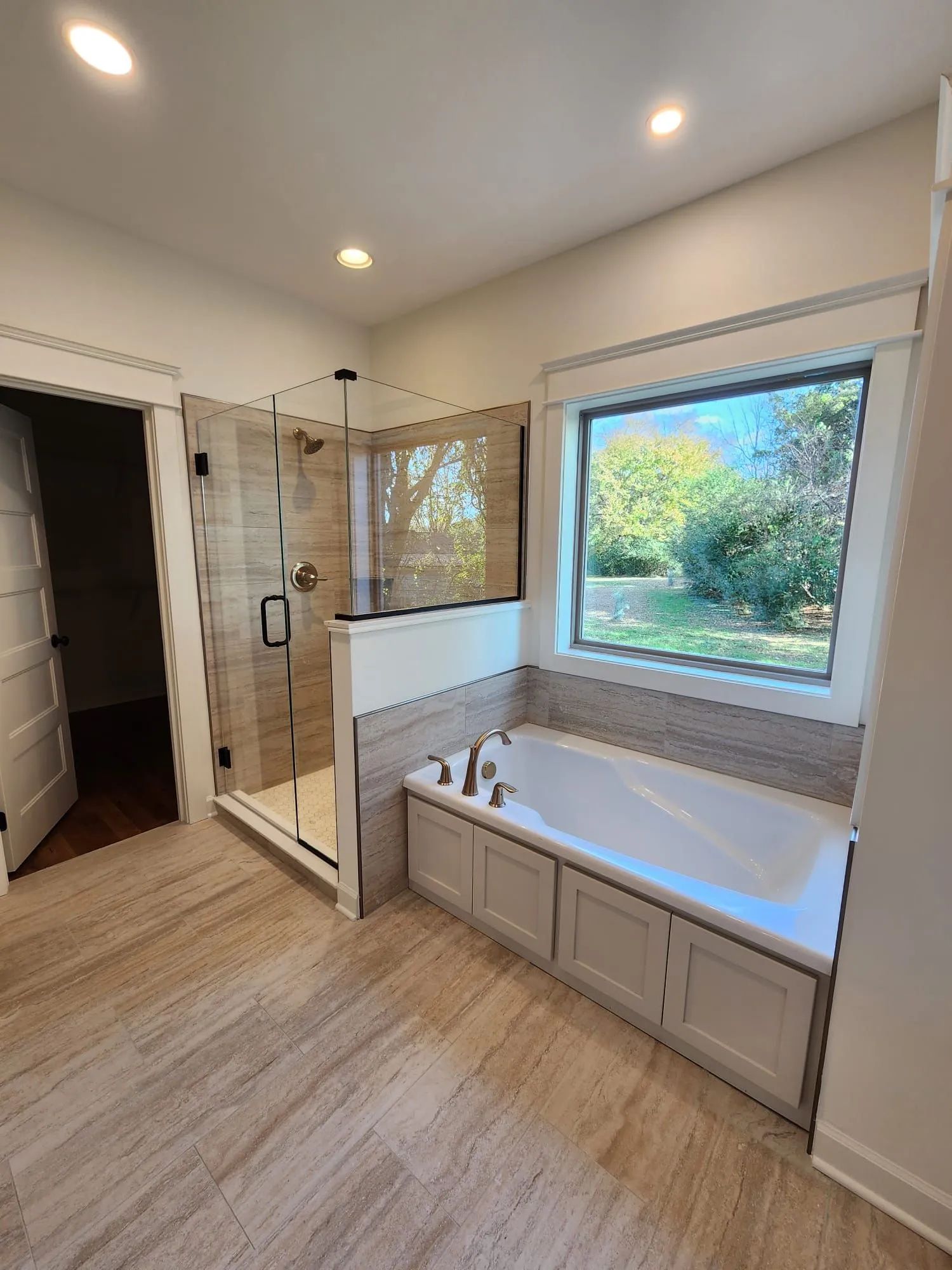 Modern bathroom with glass shower, bathtub, and large window overlooking greenery