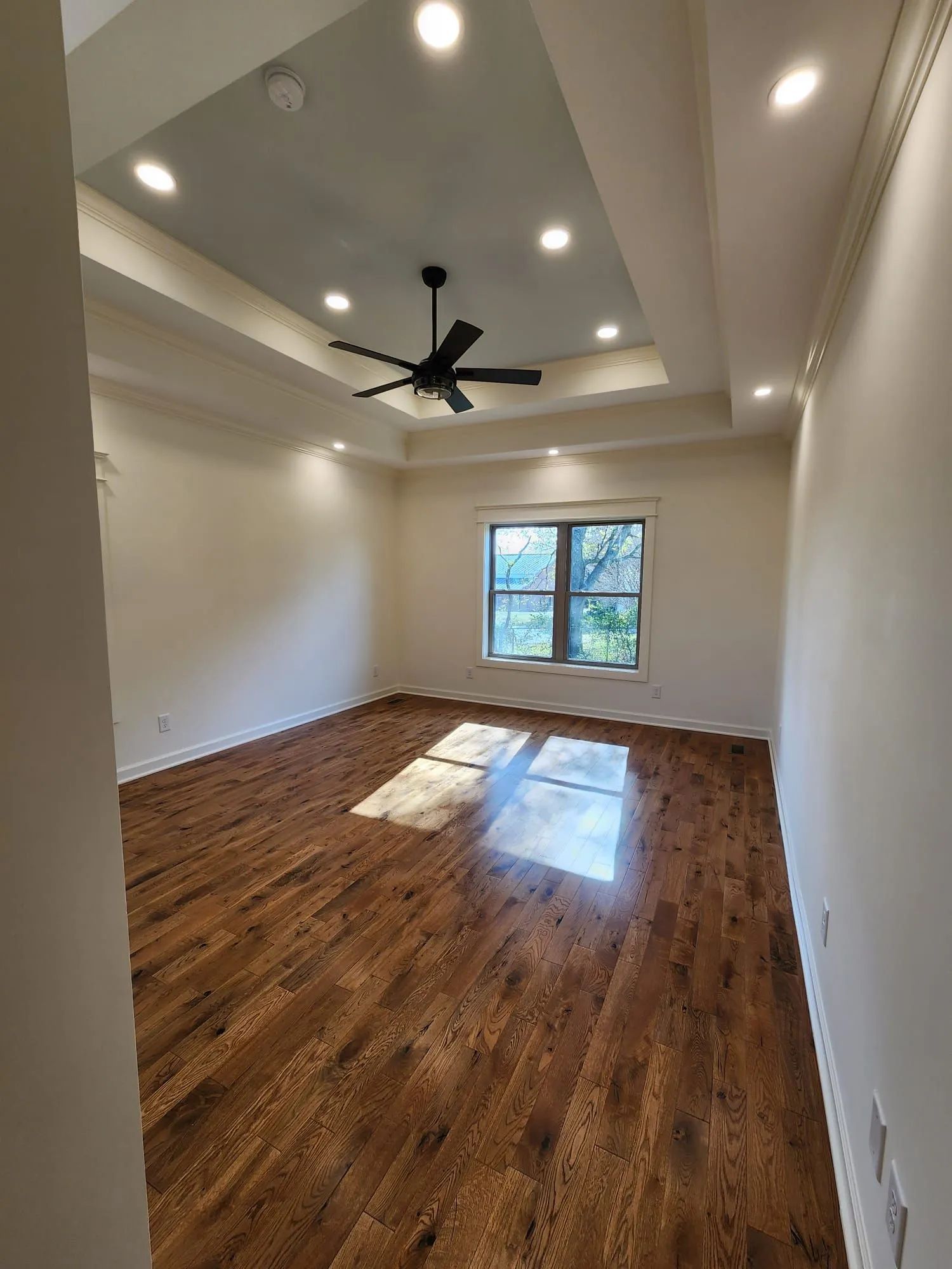 Empty living room with wood floors, tray ceiling, ceiling fan, and sunlight from a window.