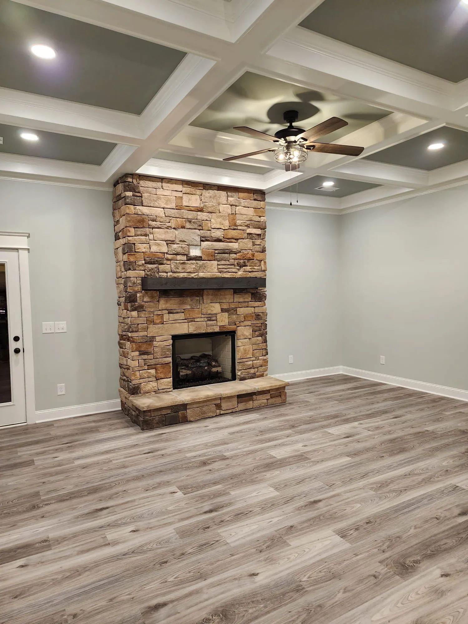 Empty living room with stone fireplace, coffered ceiling, ceiling fan, and gray wood-look floors