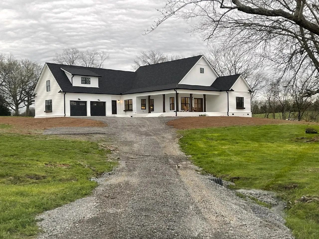 White farmhouse with black roof at the end of a gravel driveway in a grassy rural yard