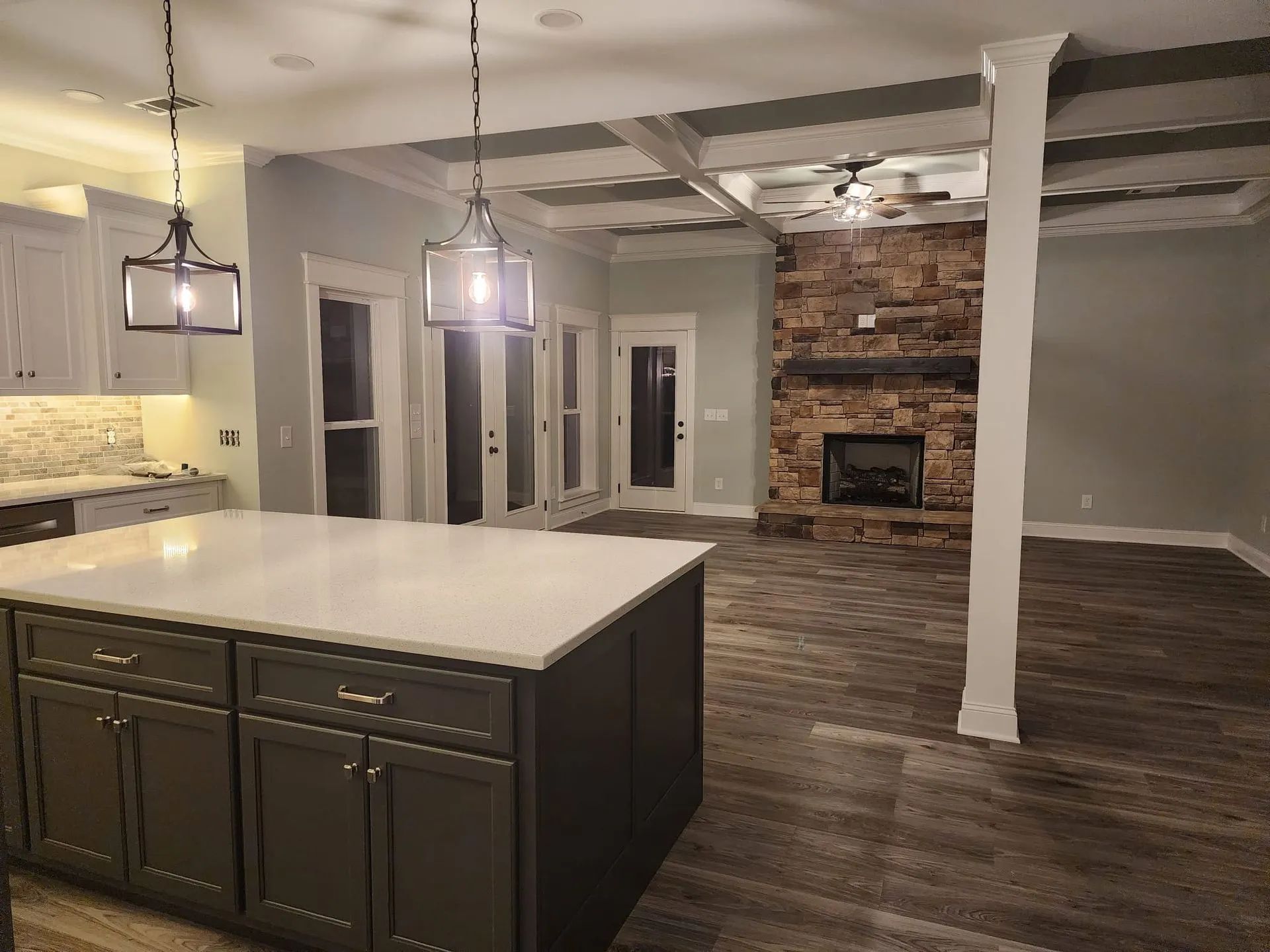 Modern kitchen with large island opening to a living room, featuring pendant lights and a brick fireplace.