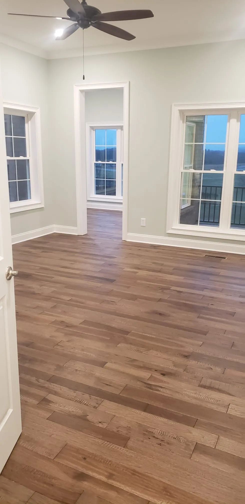 Empty room with hardwood floors, ceiling fan, white walls, and tall windows with city views.