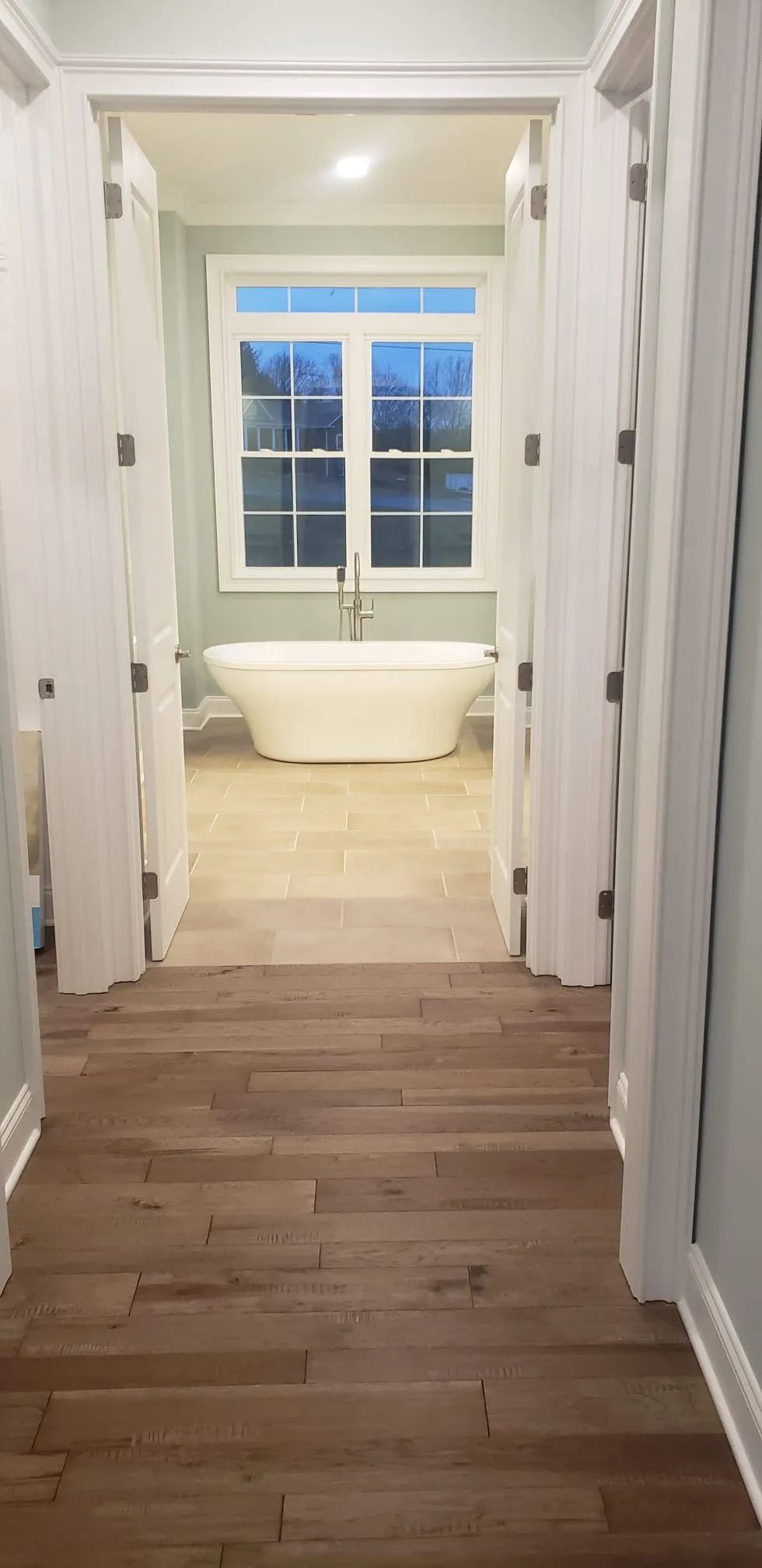 Hallway with wood floors leading to a bright bathroom with a freestanding tub by a window