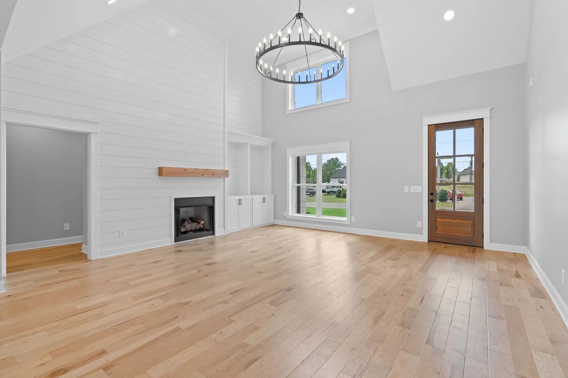 Bright empty living room with hardwood floors, white brick fireplace, chandelier, and large windows.