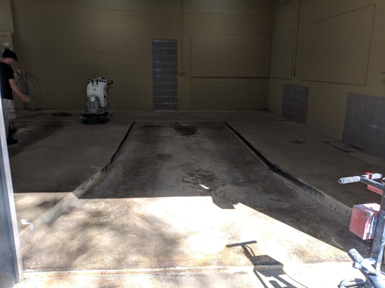 Concrete floor with depressions. Man stands near wall, floor polisher in the center. Sunlight enters the opening.