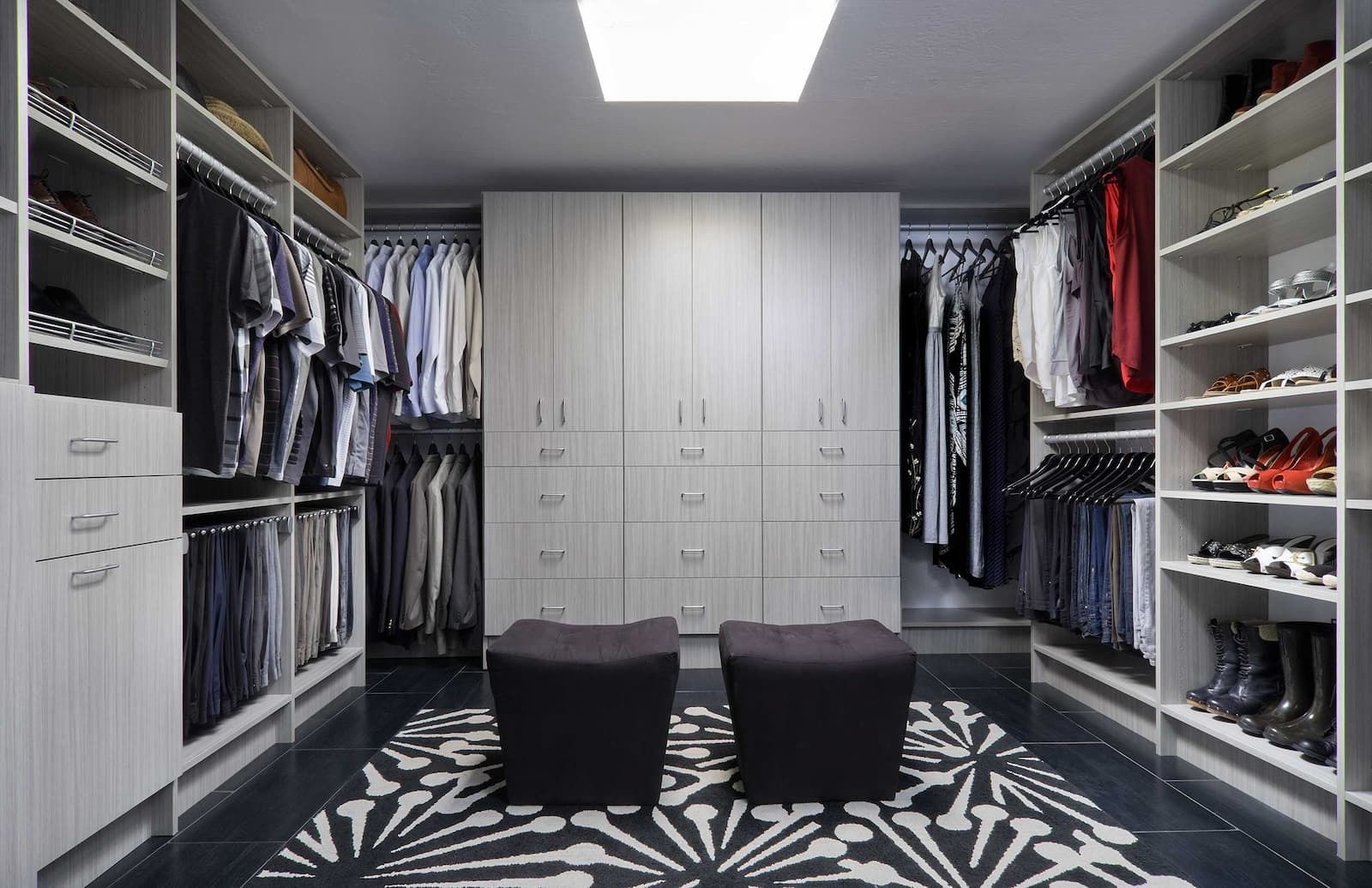 Walk-in closet with gray shelving, drawers, hanging clothes, shoes. Two stools on a patterned rug.