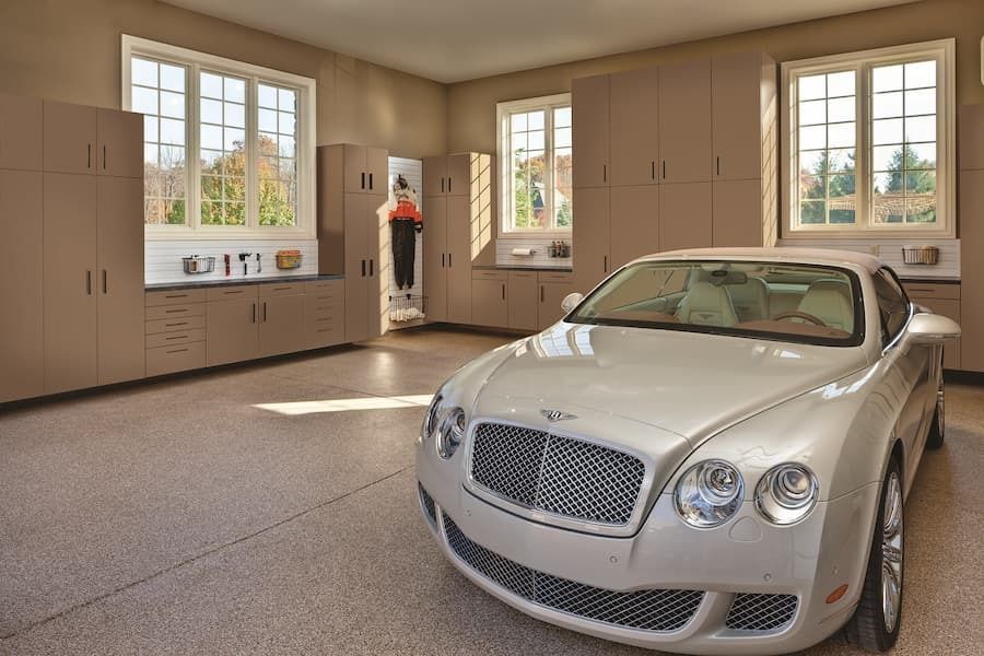 Silver Bentley parked in a tan garage with storage cabinets and windows.