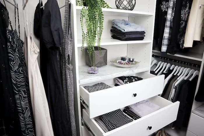 White closet organizer with drawers, holding folded clothes and a potted plant, surrounded by hanging garments.