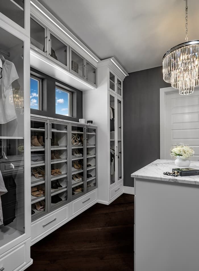 Walk-in closet with white cabinets, glass-front shoe shelves, dark wood floor, and chandelier.
