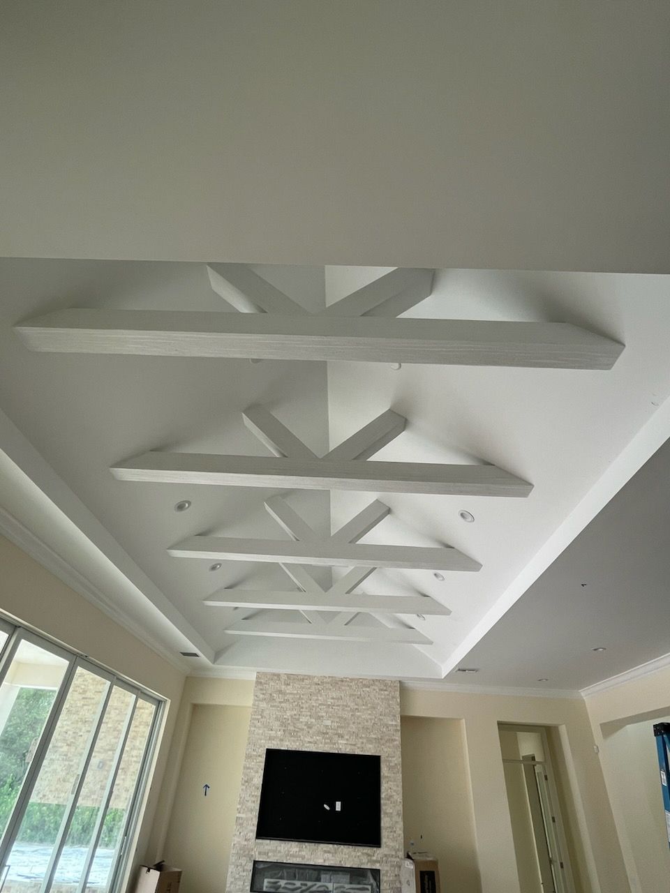 Modern living room ceiling with white beams, a stone fireplace, and large windows letting in daylight.