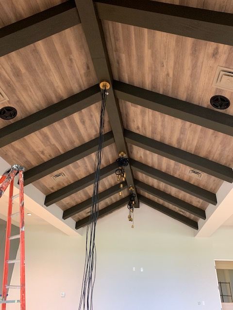 Vaulted ceiling with exposed dark beams, wood panels, and hanging electrical wires in a room under construction.