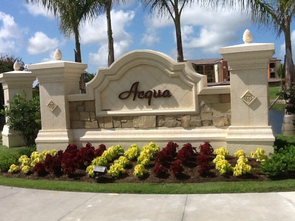 Aqua entrance sign with cream stone columns and colorful flower beds under palm trees