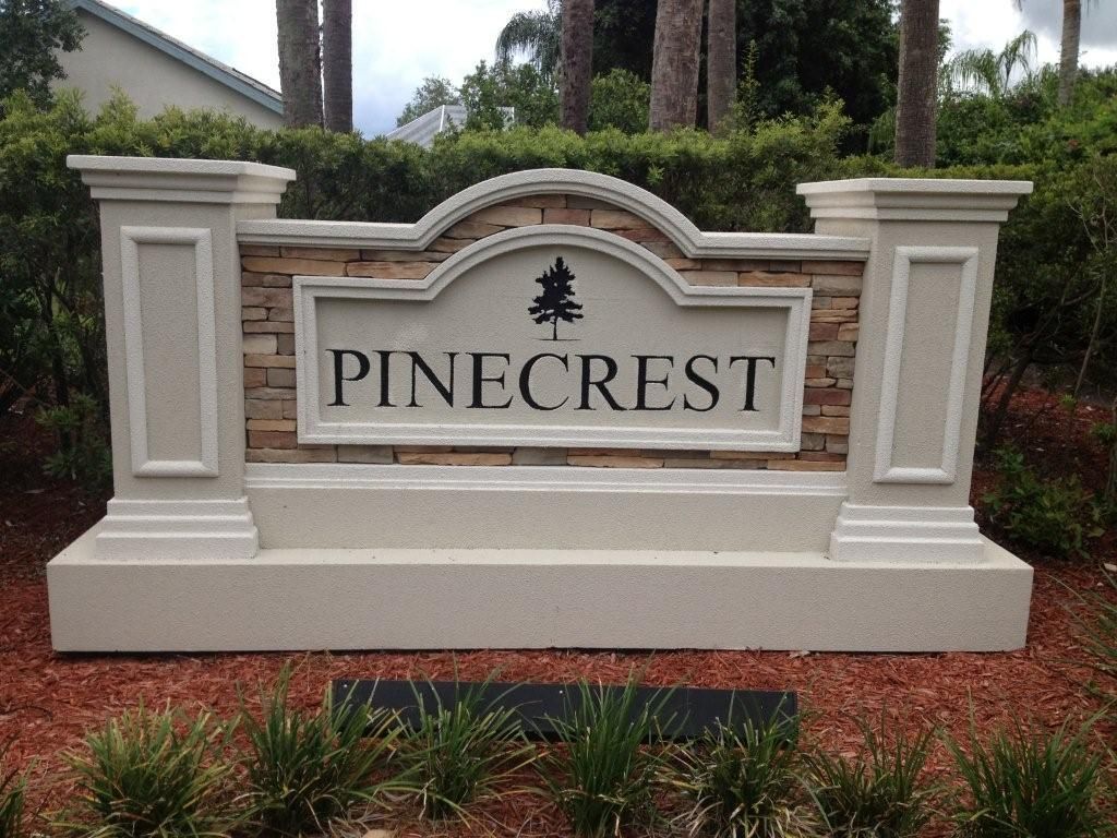 Pinecrest community entrance sign in beige stone with a landscaped base and trees behind it