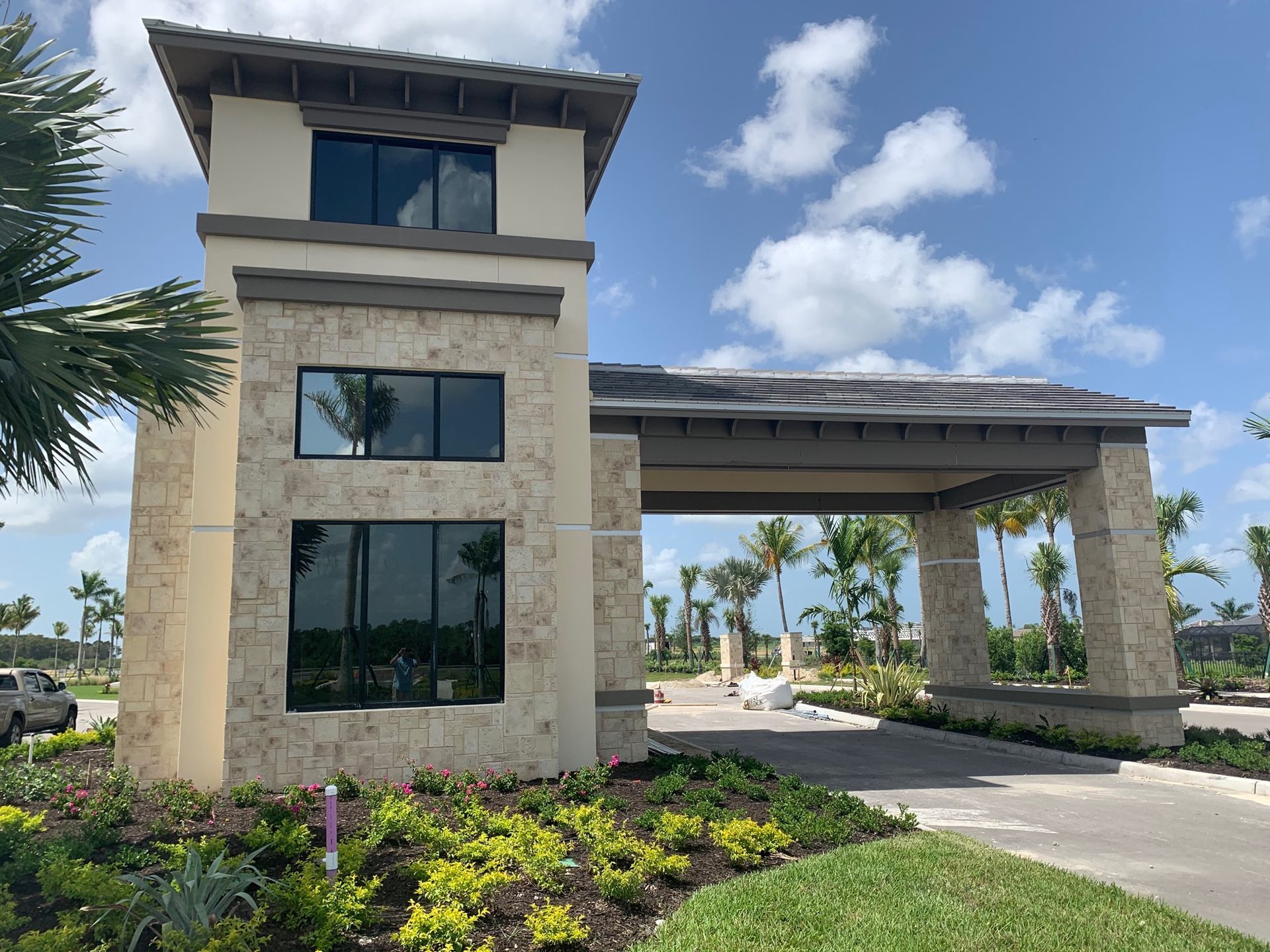 Modern building entrance with stone columns, dark windows, covered driveway, and landscaped greenery under a blue sky.