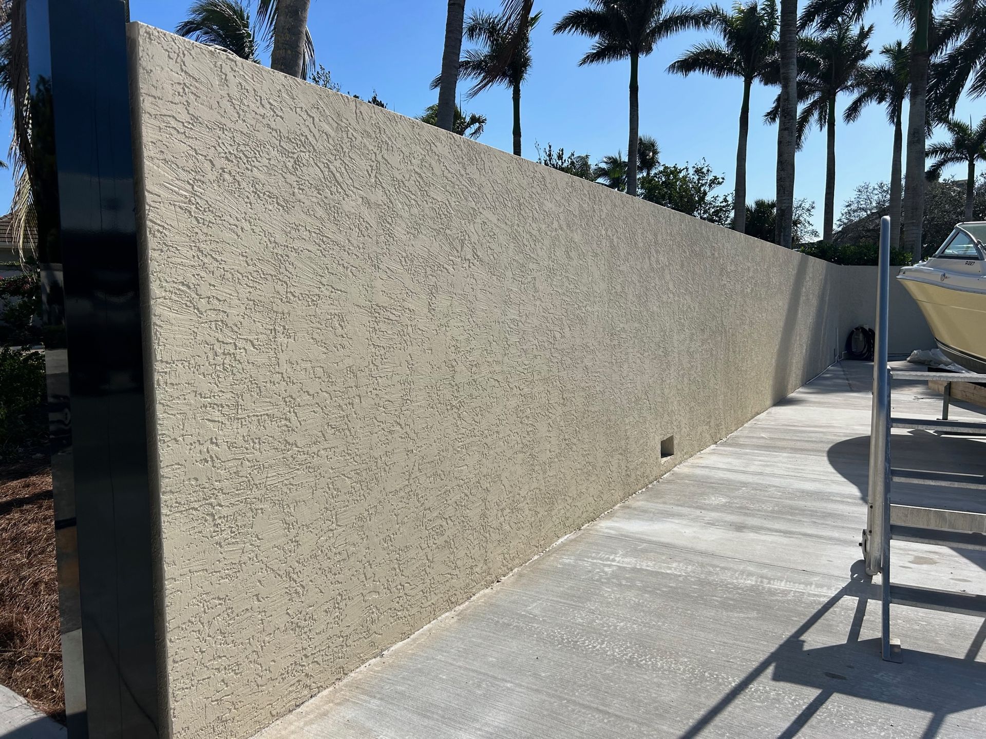 Long beige stucco wall beside a concrete walkway, with a metal scaffold and palm trees in the background.