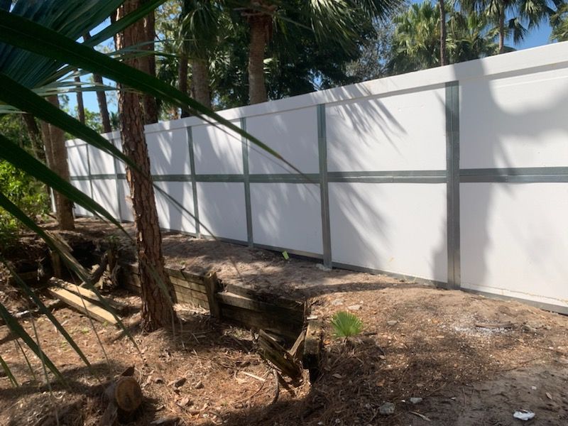 White privacy fence along a shaded dirt path with palm trees and a wooden bench in a tropical yard.