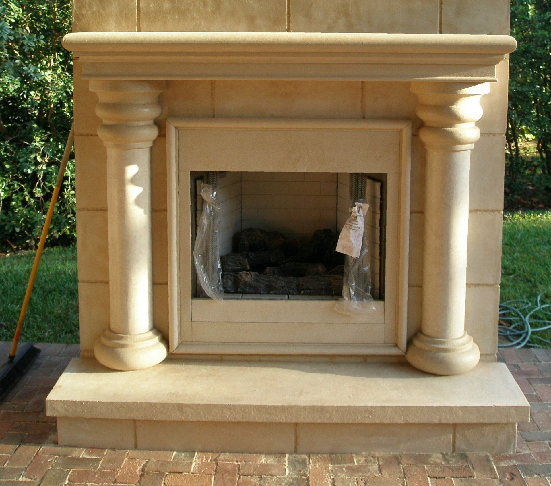 Cream stone fireplace with ornate columns and a glass front on a brick patio.