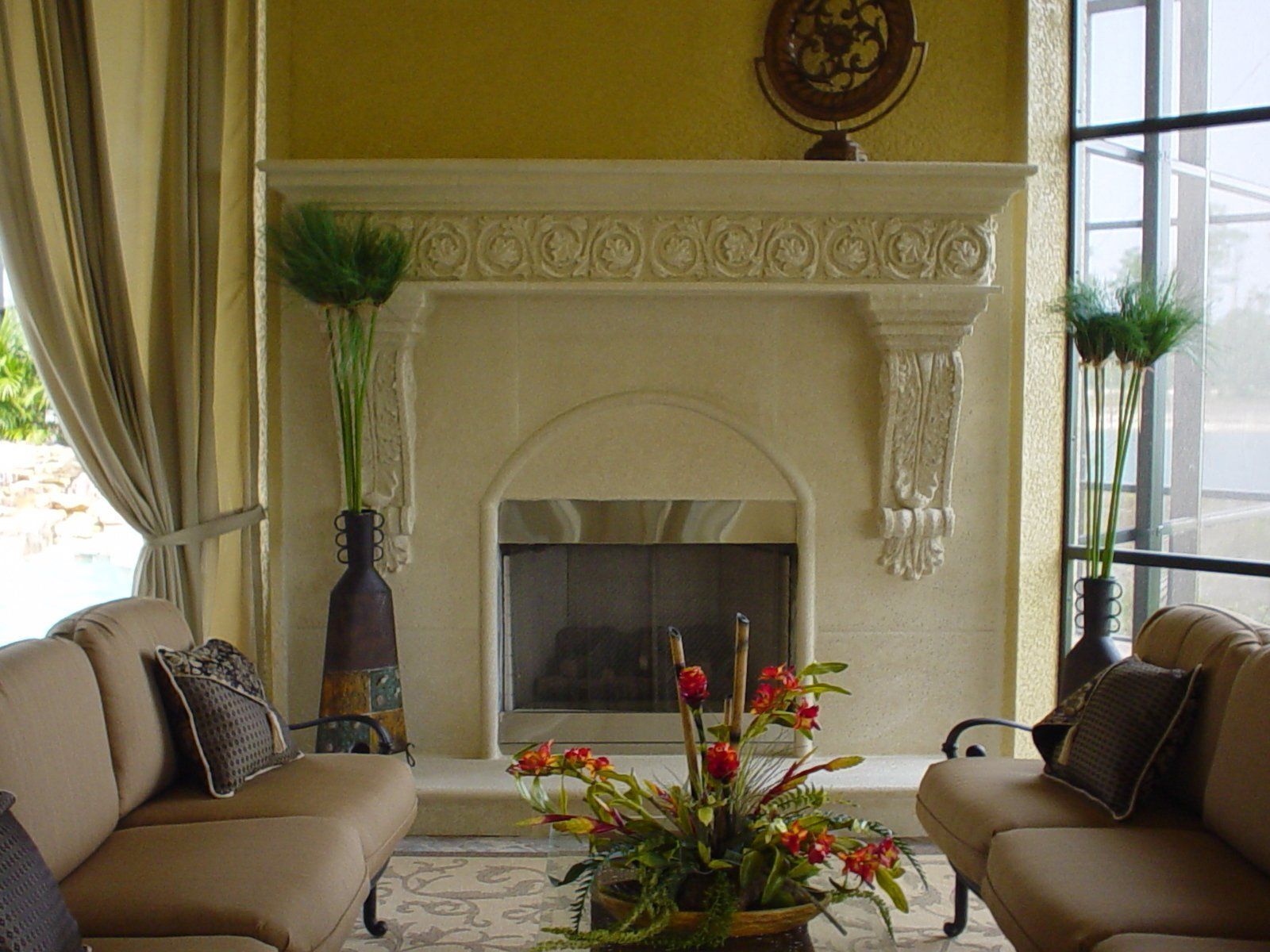 Elegant living room with cream fireplace, beige sofas, tall windows, and a floral coffee table centerpiece