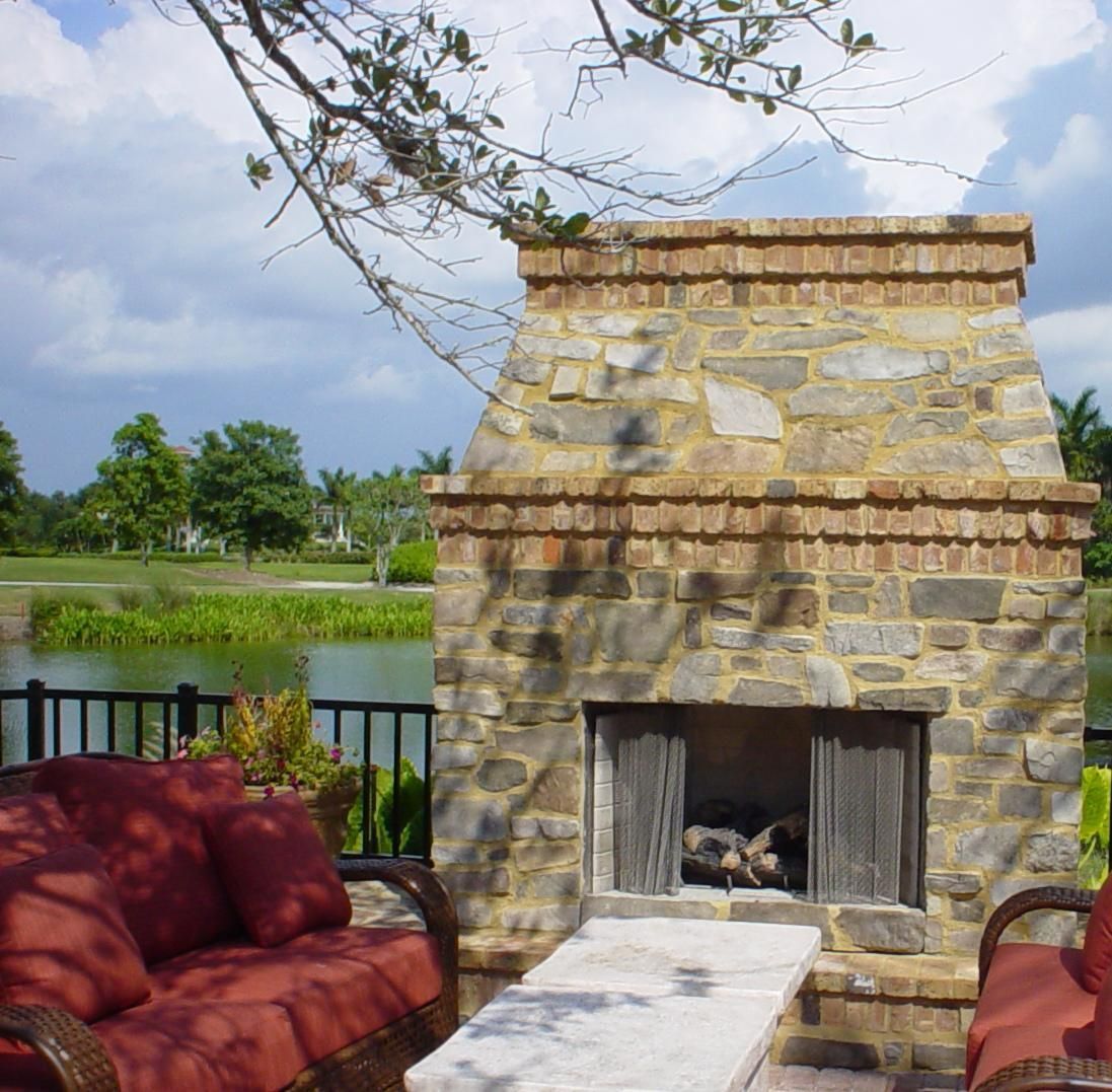Stone outdoor fireplace with red cushioned seating beside a lake and grassy yard