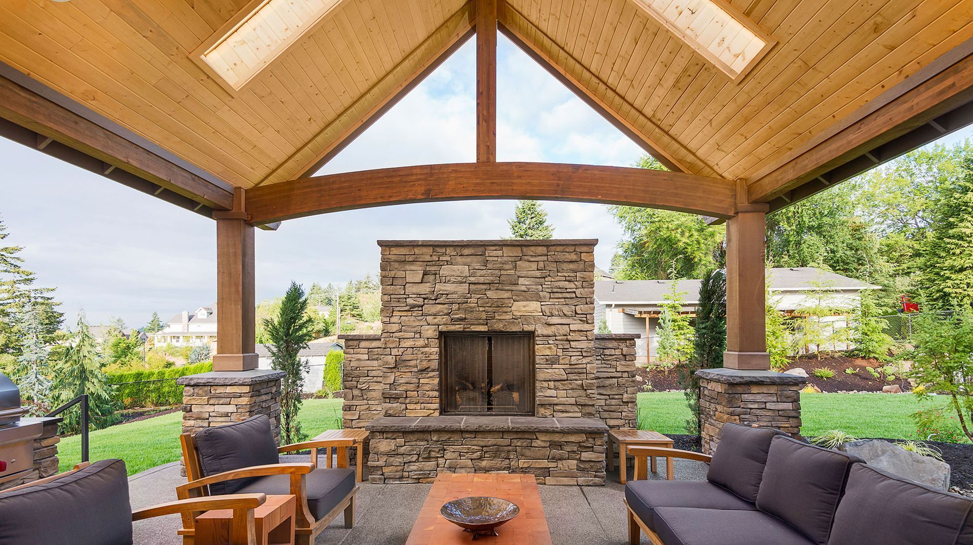 Covered outdoor patio with wood ceiling, stone fireplace, and seating overlooking a garden