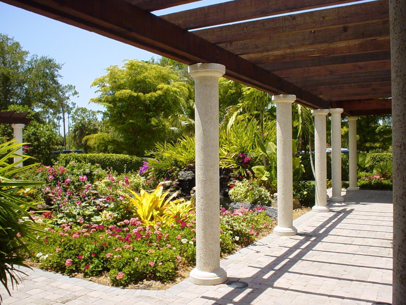 Covered garden patio with white columns, stone walkway, and colorful flowers in bright sunlight