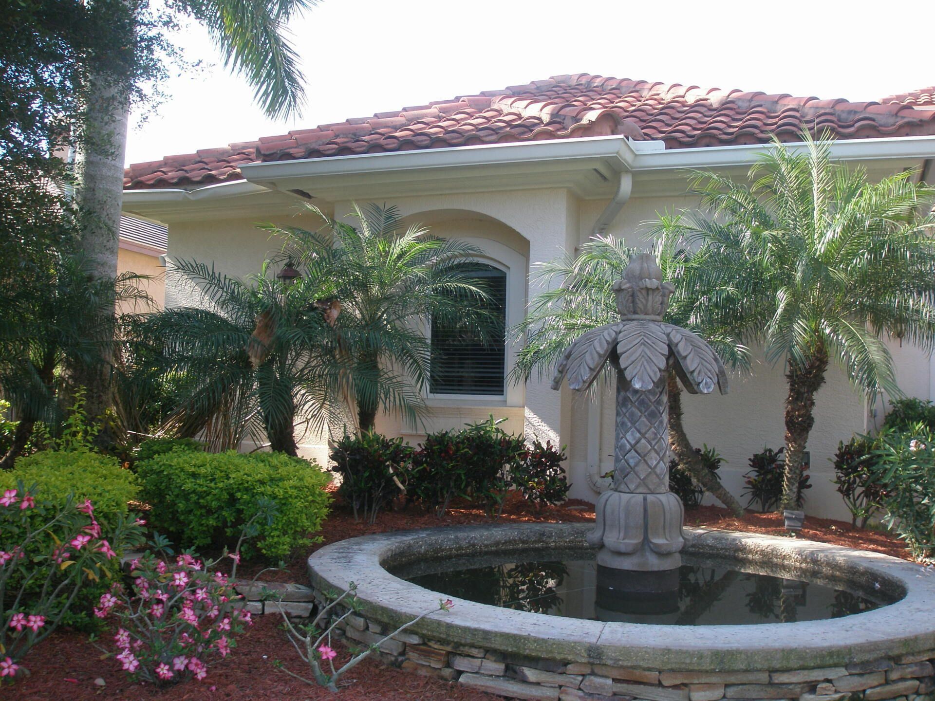 Courtyard garden with a small fountain statue, palm trees, pink flowers, and a white stucco house