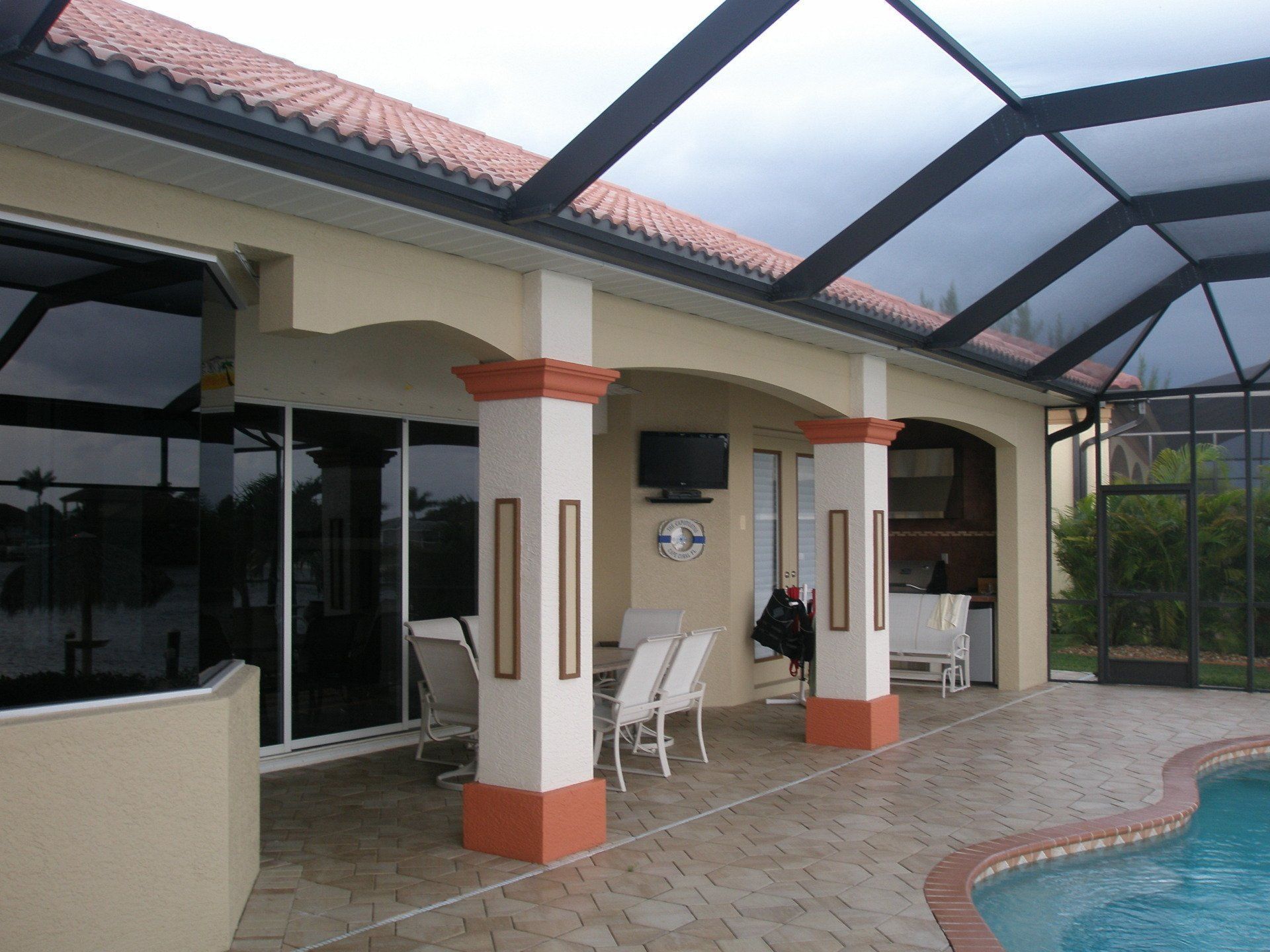 Covered patio beside a swimming pool with white chairs, glass doors, and a tiled roof.