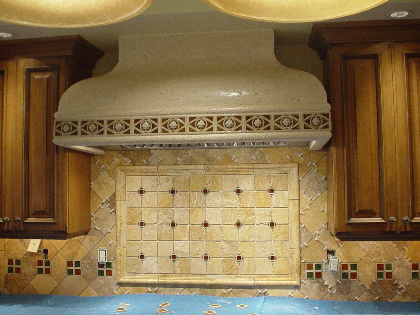 Kitchen backsplash with beige tile, decorative vent hood, and wooden cabinets on either side