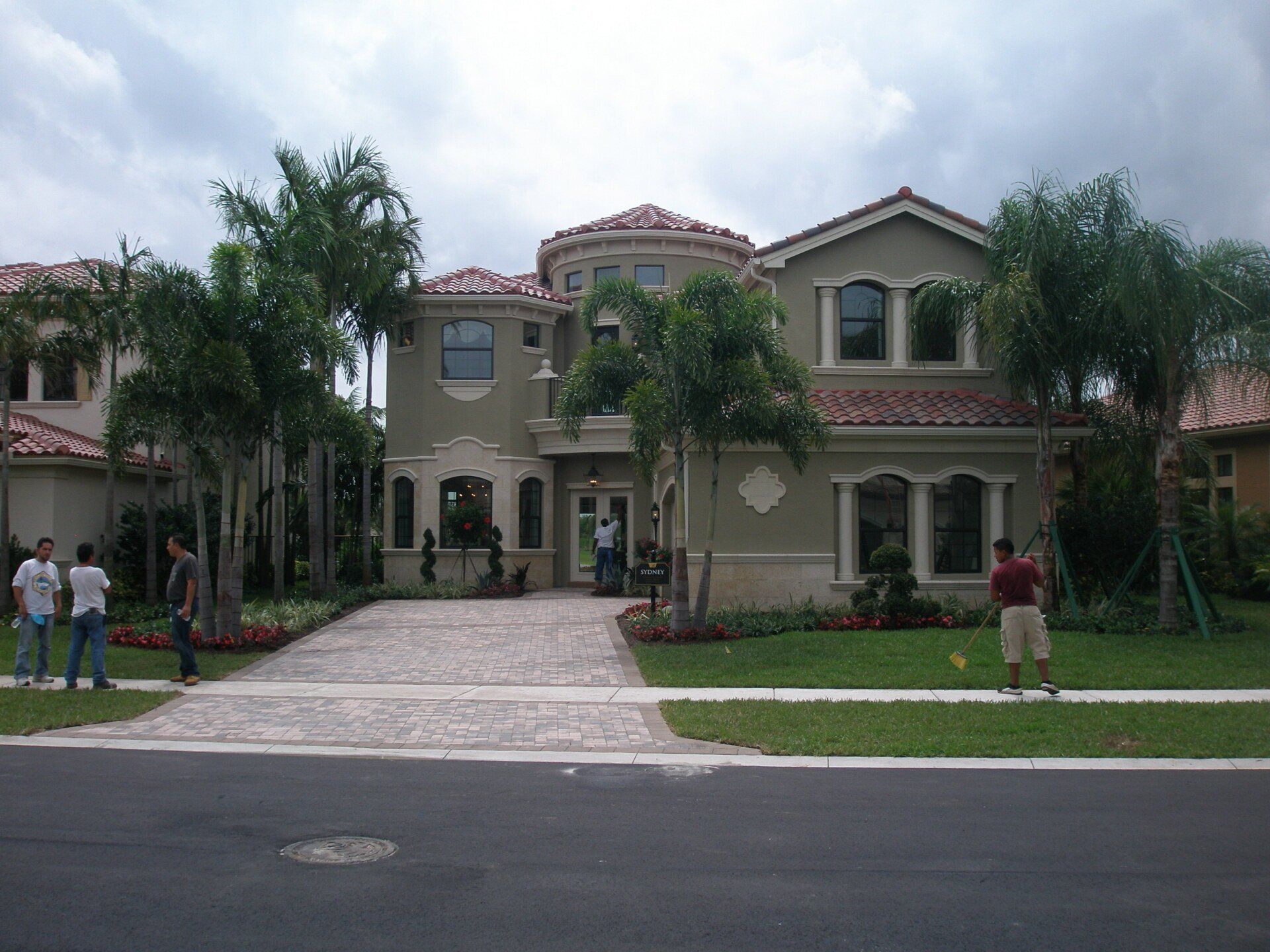Large house with a tiled roof, front lawn, driveway, and a few people standing outside under cloudy skies