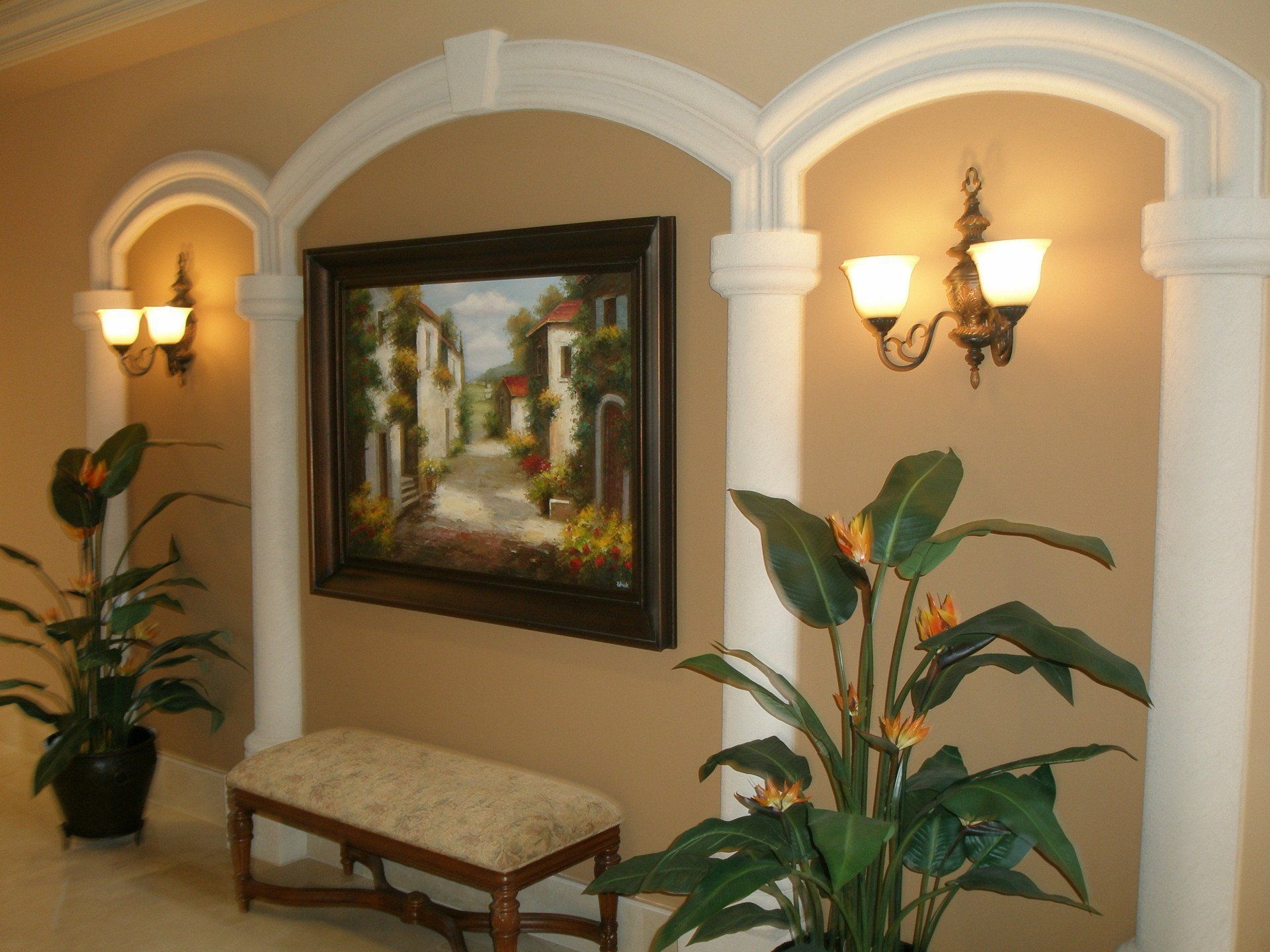 Elegant hallway with arched alcoves, wall sconces, framed art, bench, and potted plants