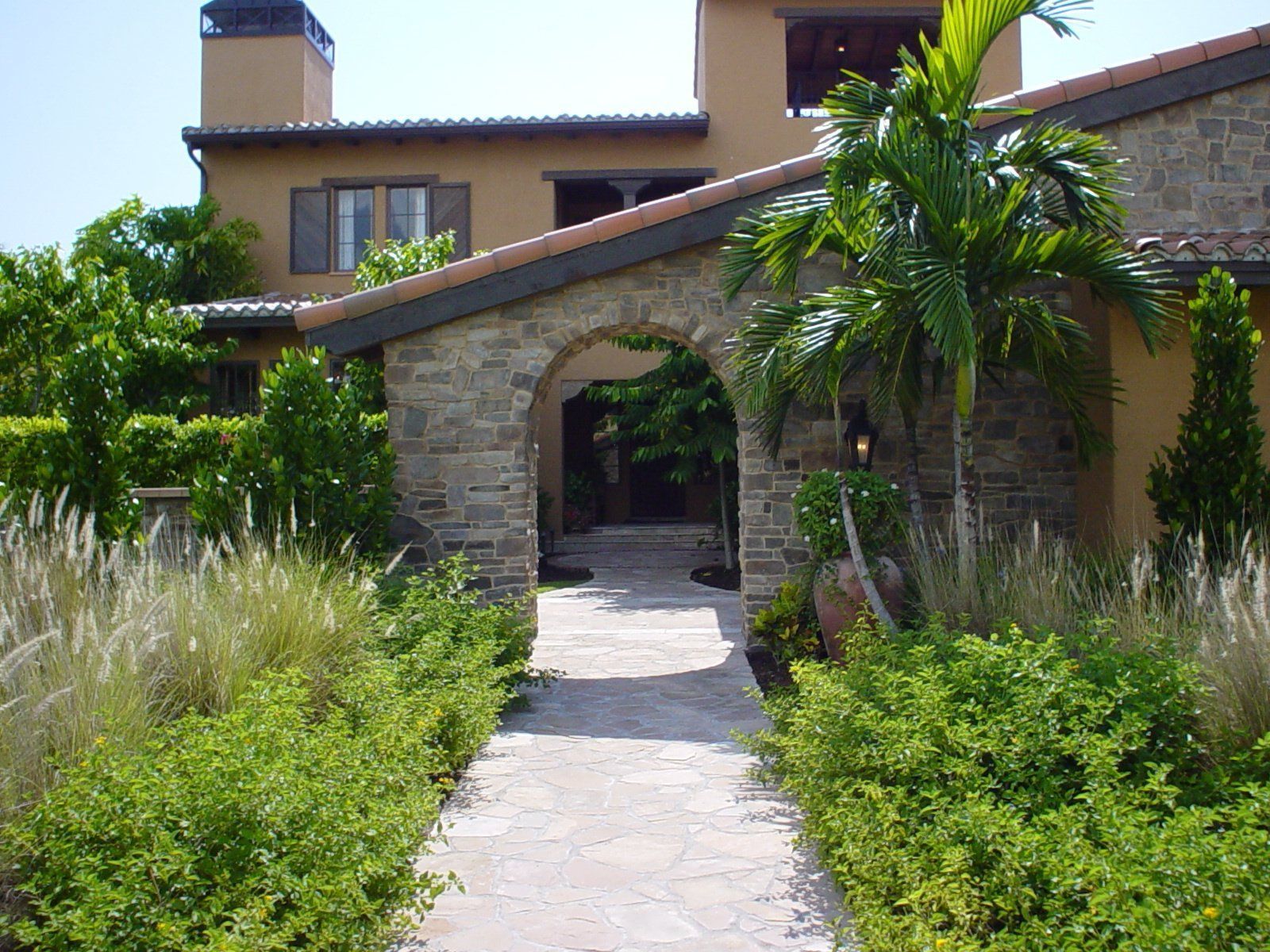 Stone-arched garden walkway leading to a tan house with palm trees and lush greenery