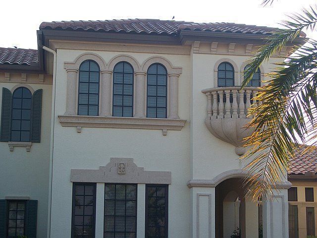 Mediterranean-style stucco house facade with arched windows and a small balcony, partly shaded by palm leaves