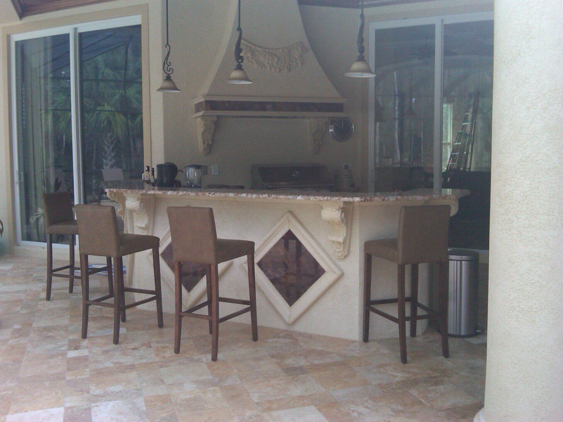 Outdoor kitchen bar with high stools, marble counter, grill hood, and sliding glass doors.