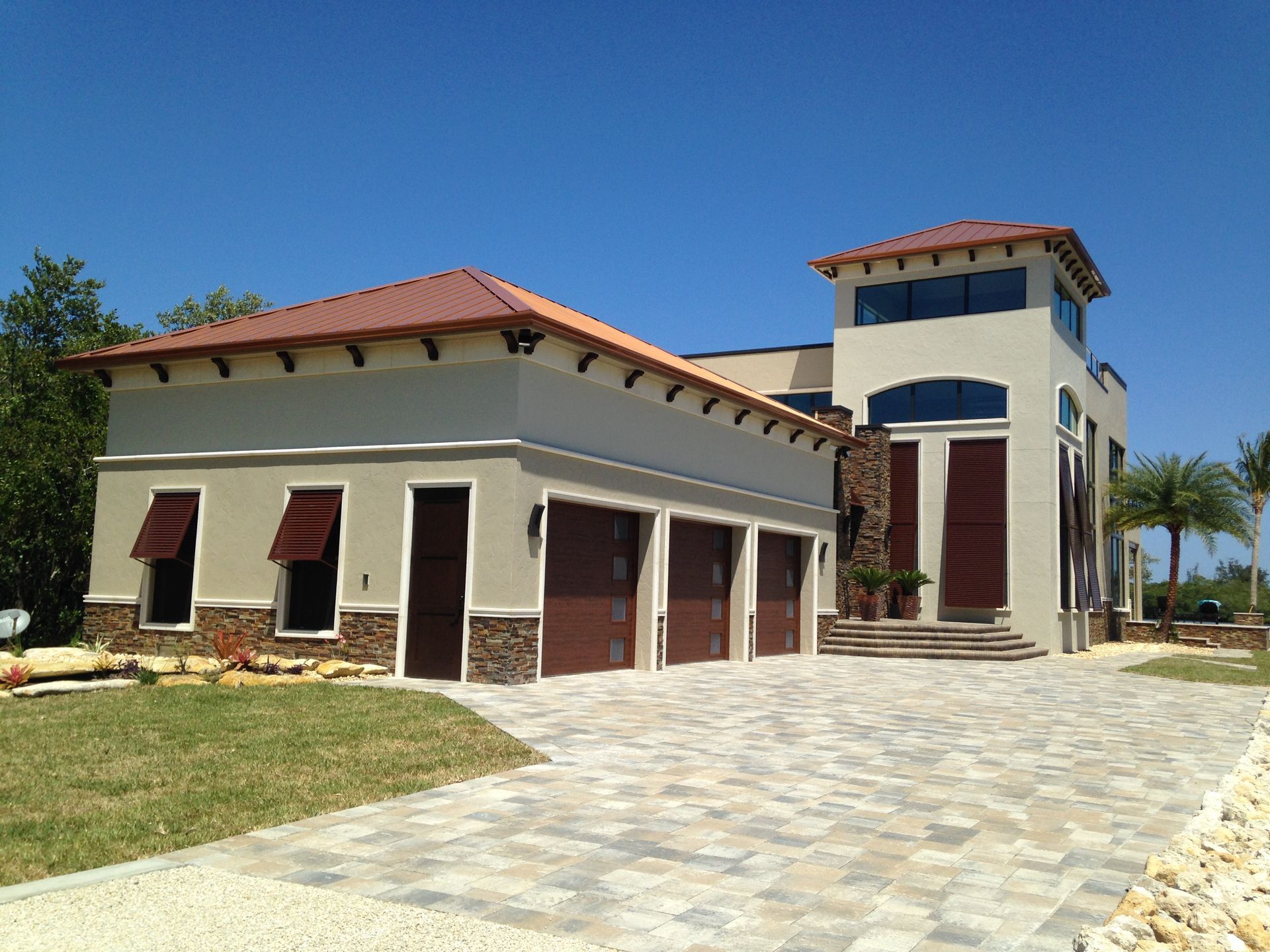 Two-story beige house with red tile roof, arched entry, and stone driveway under a clear blue sky.