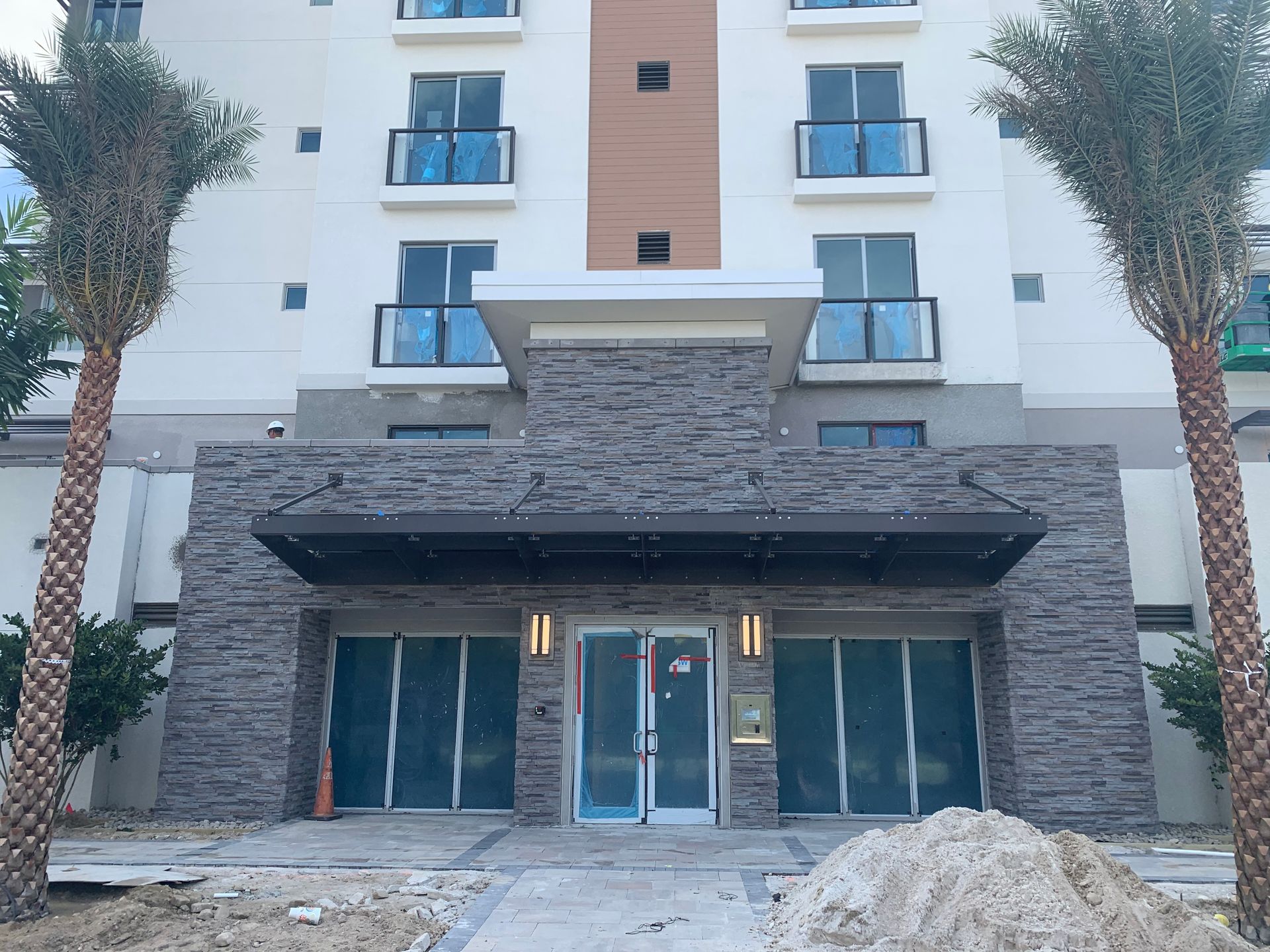 Modern building entrance with stone facade, glass doors, balconies, and palm trees on both sides