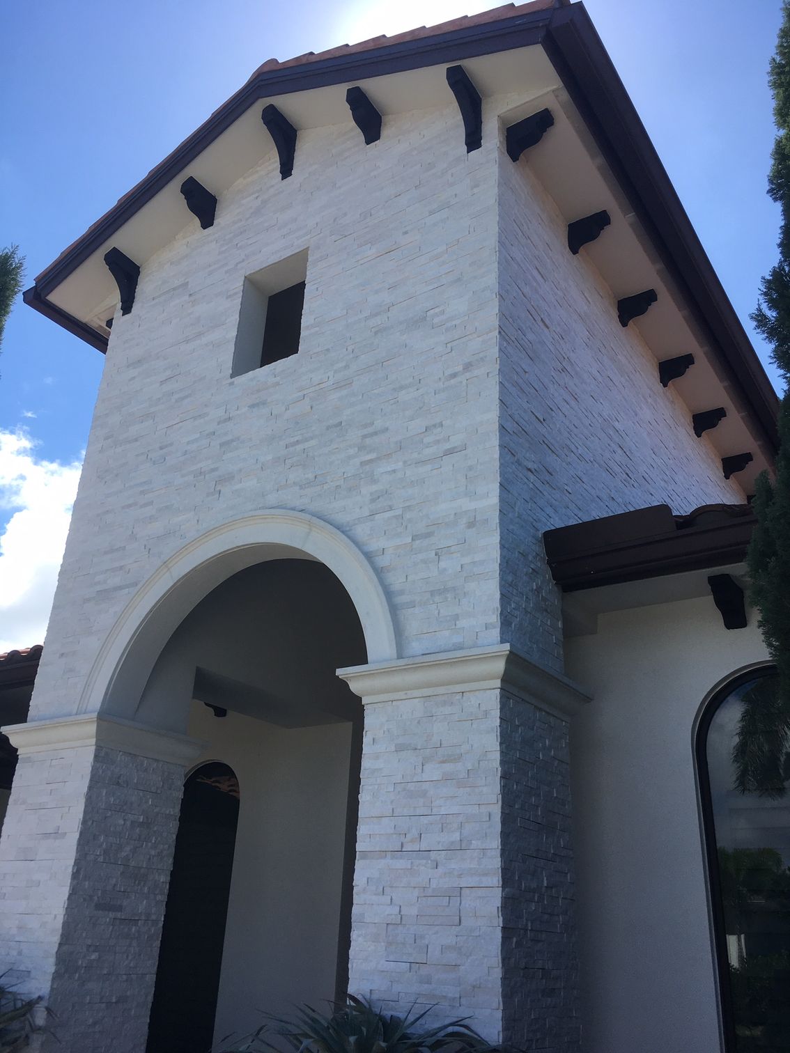 White stucco building with arched entryway, dark roof tiles, and exposed beams against a blue sky.
