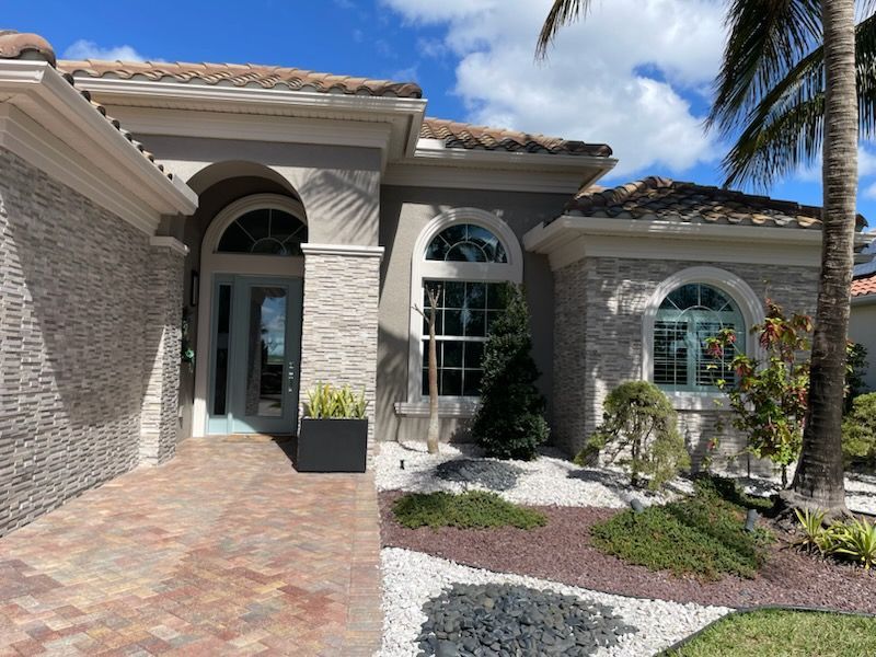 Front entrance of a stucco house with arched windows, brick walkway, and palm trees under a blue sky.