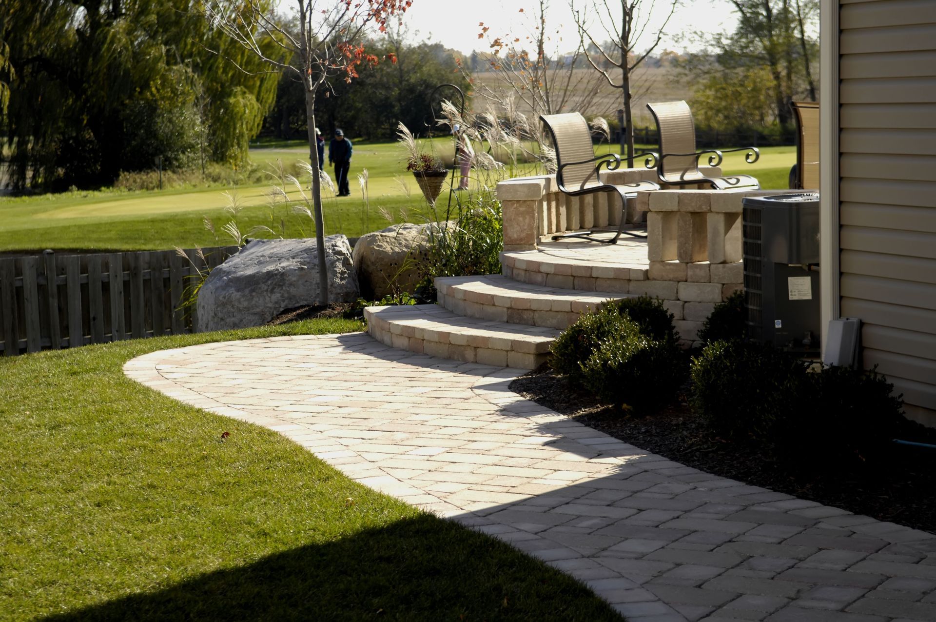 A paved patio and walkway with stone steps leading to a seating area. Lawn in foreground.