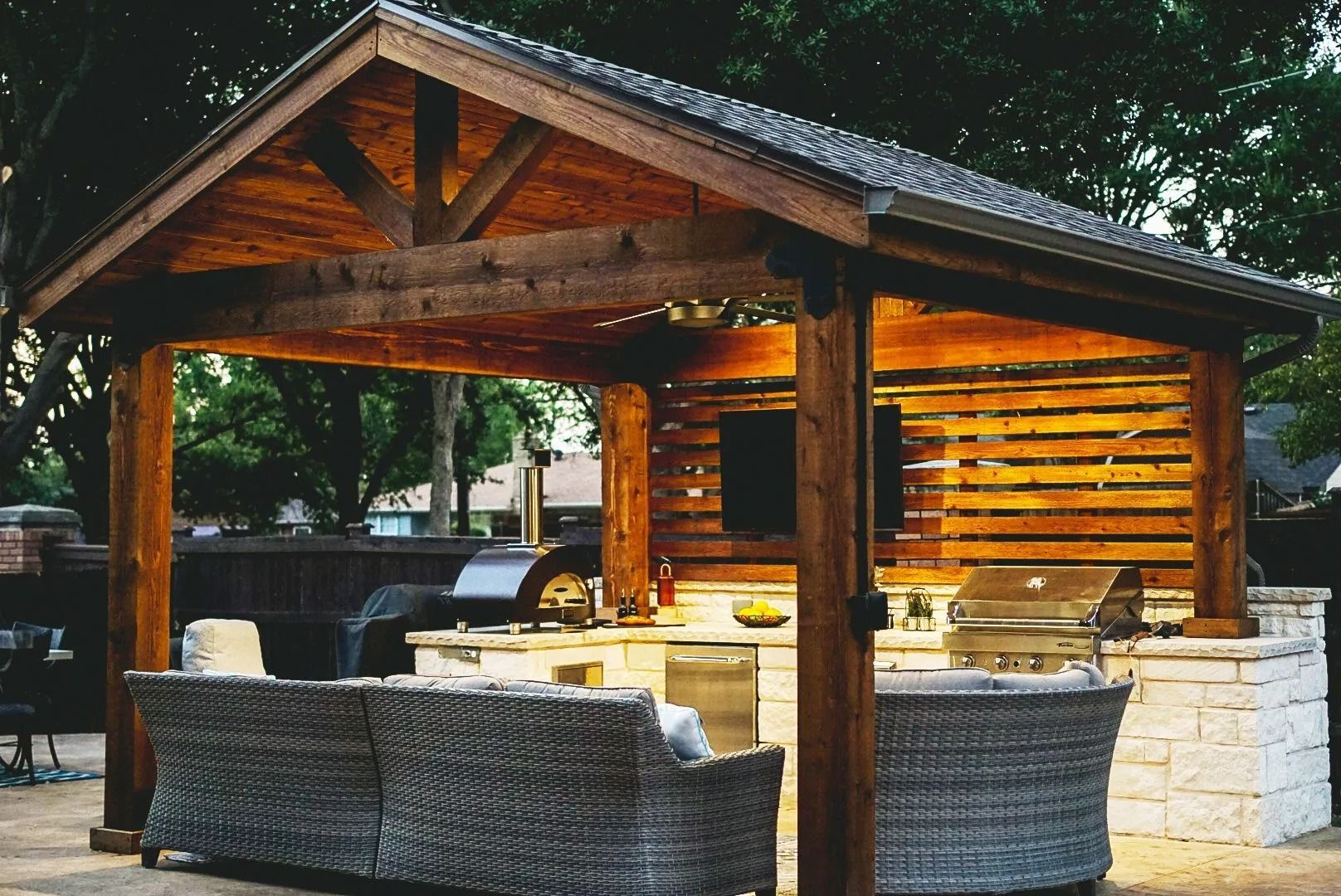 Wooden outdoor gazebo with stone walls, wicker seating, and a built-in grill at dusk